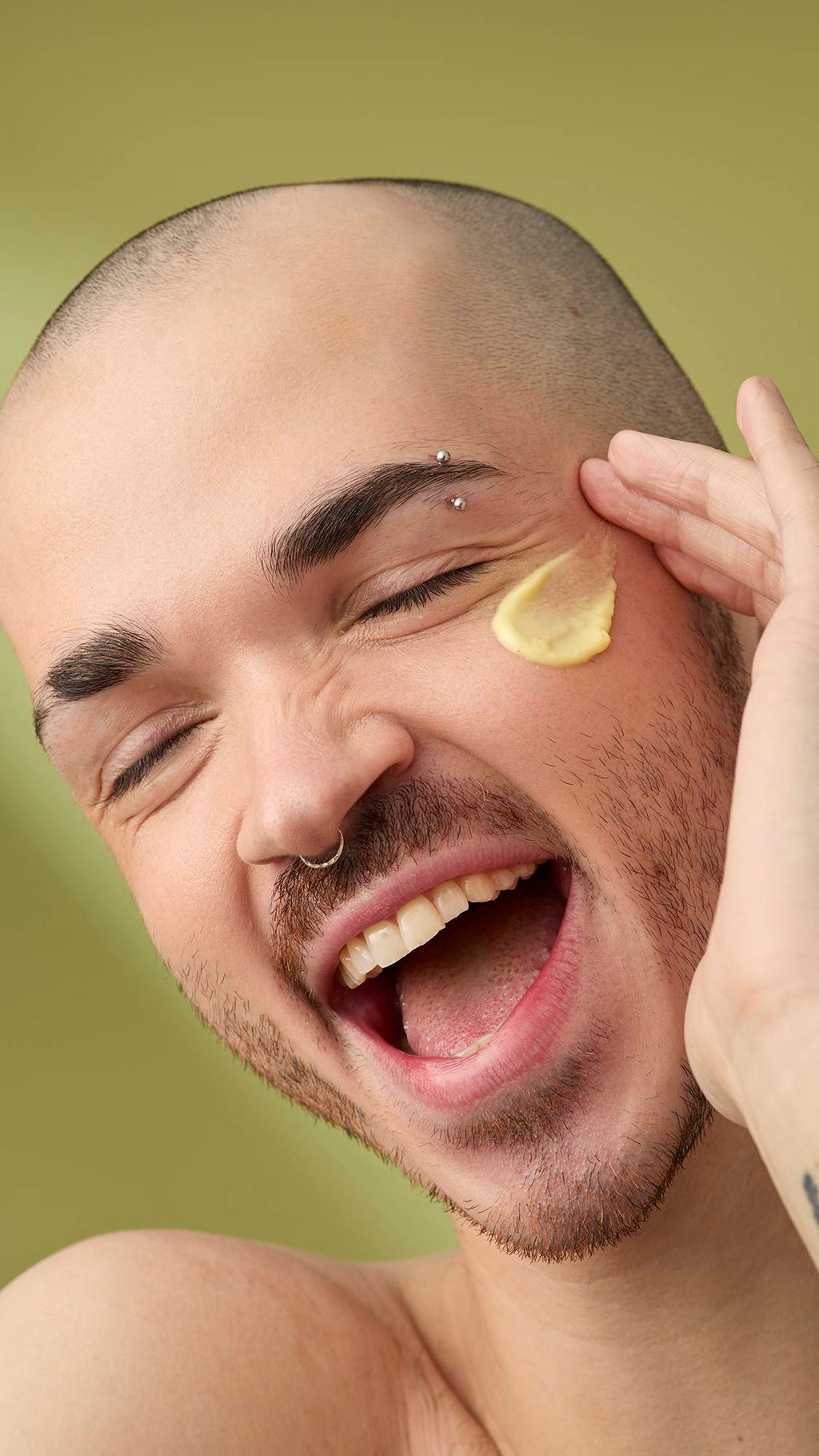 A person with a shaved head and facial piercings laughs as they apply a dab of Acai And Sacha Inchi facial cleanser to their cheek. They have an eyebrow bar and nose ring, and are shown against a solid olive-green background.