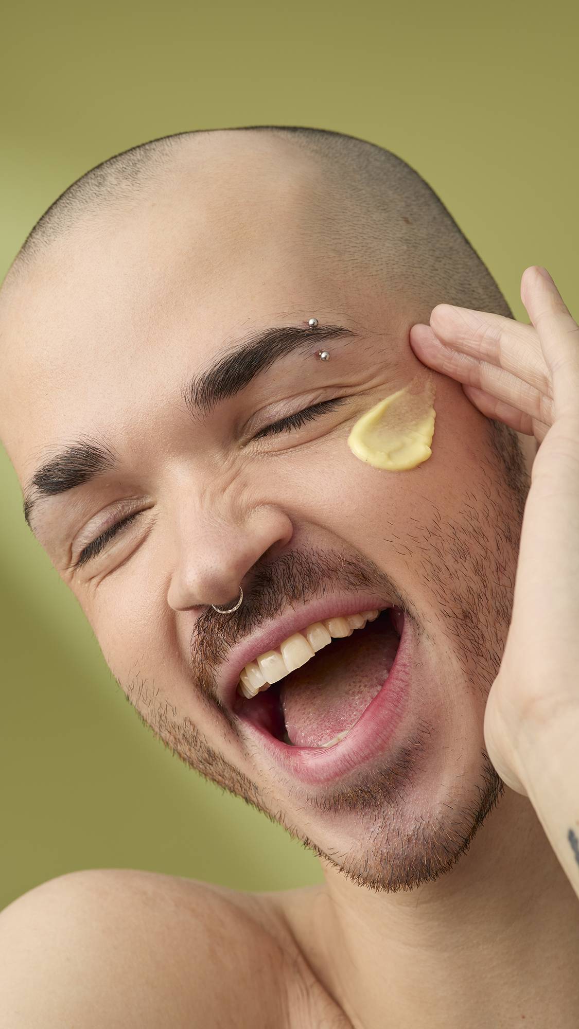 A person with a shaved head and facial piercings laughs as they apply a dab of Acai And Sacha Inchi facial cleanser to their cheek. They have an eyebrow bar and nose ring, and are shown against a solid olive-green background.