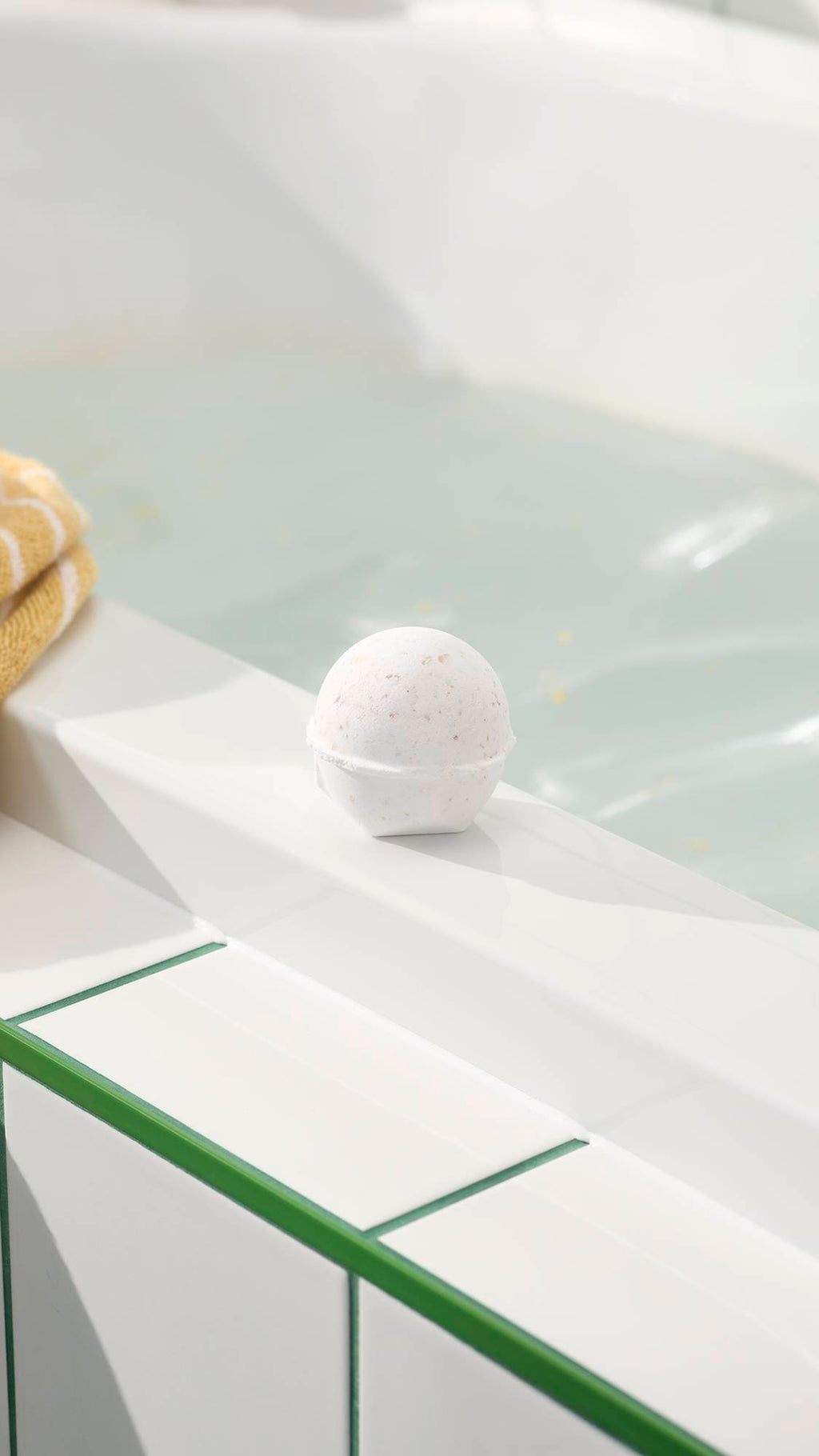 An aerial view of a white, spherical Butterball bath bomb dissolving on the surface of clear, light blue water. The product fizzes, creating a thin layer of white foam and releasing small, visible flecks of cocoa butter across the bath.