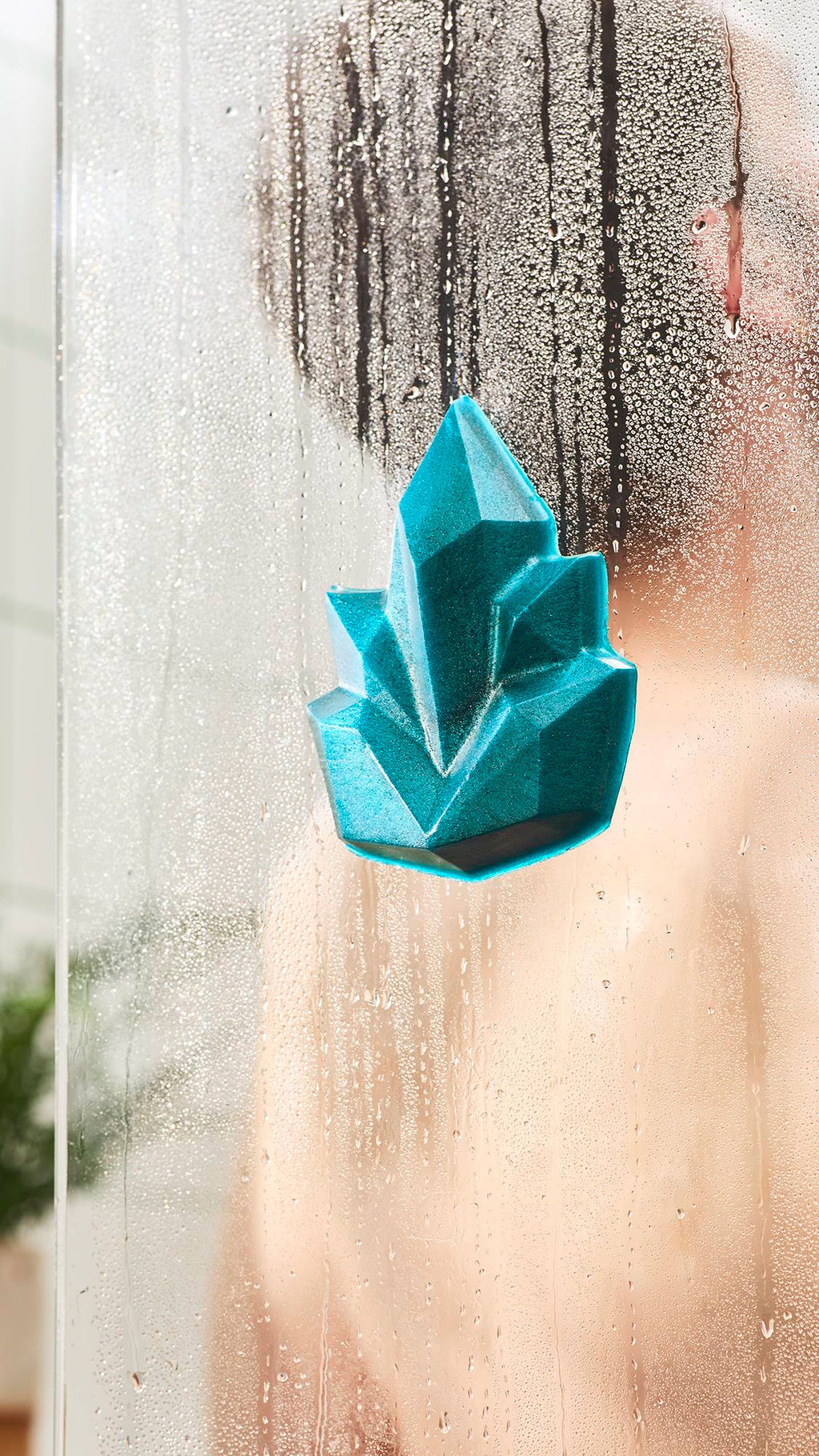 A shimmering teal, crystal-shaped shower gummy soap stuck to a steamy glass shower door. The blurry figure of a person is visible through the foggy glass covered in water droplets.