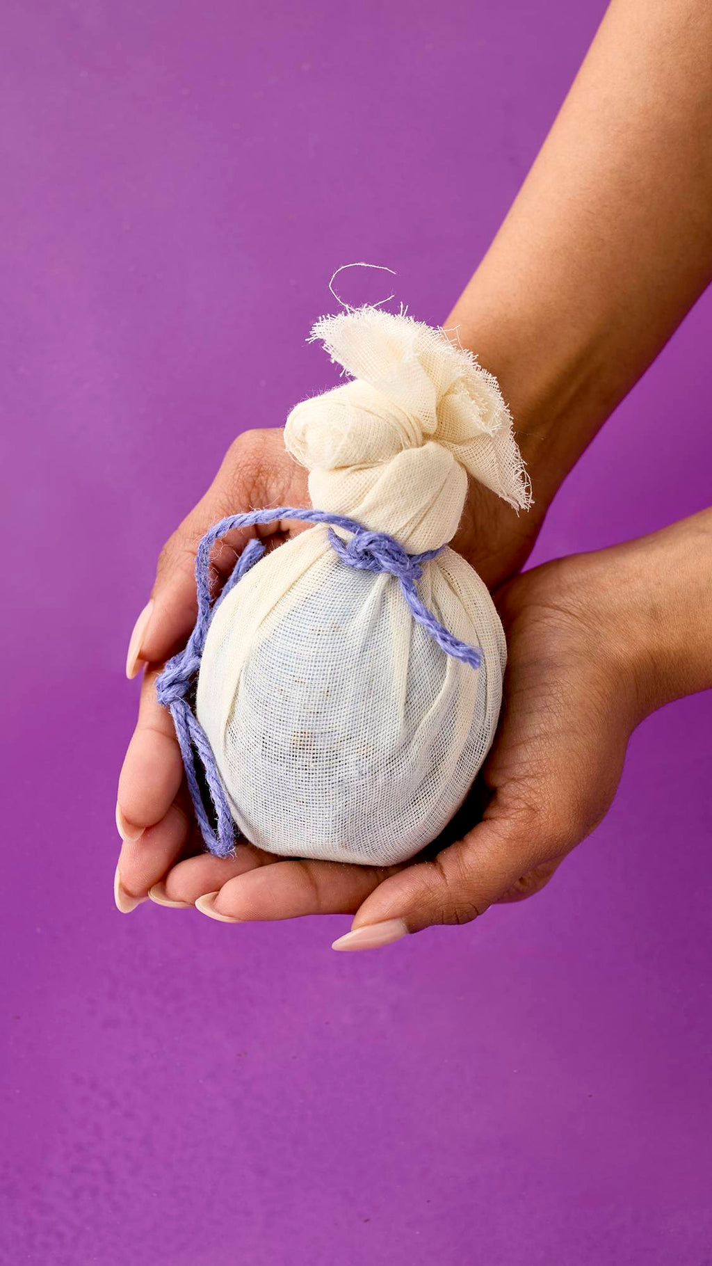 Deep Sleep. Two hands holding a spherical bath bomb wrapped in a cream-colored muslin cloth. The cloth is tied at the top with a knotted purple cord. The background is a solid, vibrant purple.