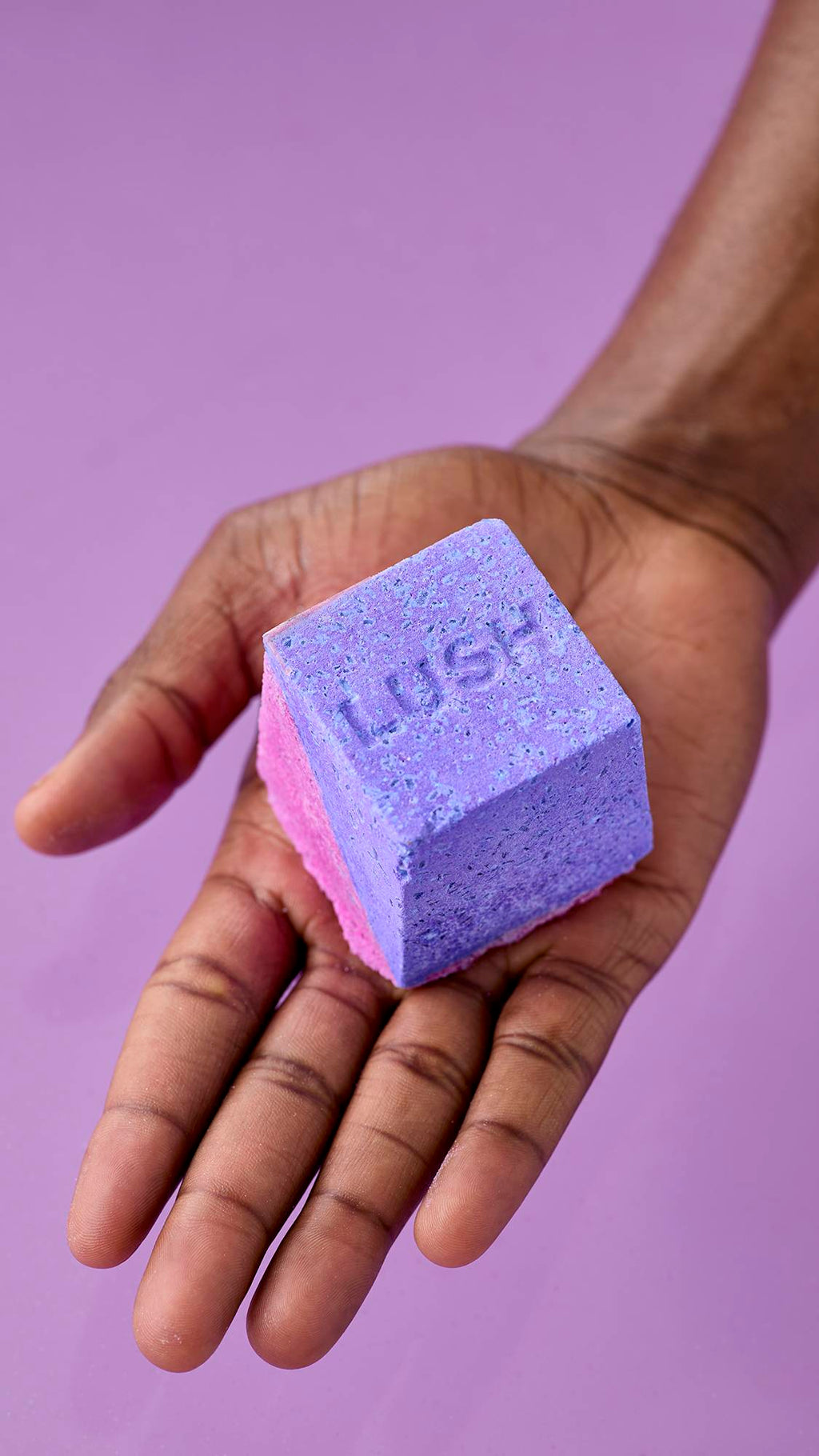 A hand holds the purple and pink Deep sleep Salt Cube bath bomb against a matching lavender background. The top surface of the cube displays a textured, embossed "LUSH" logo.