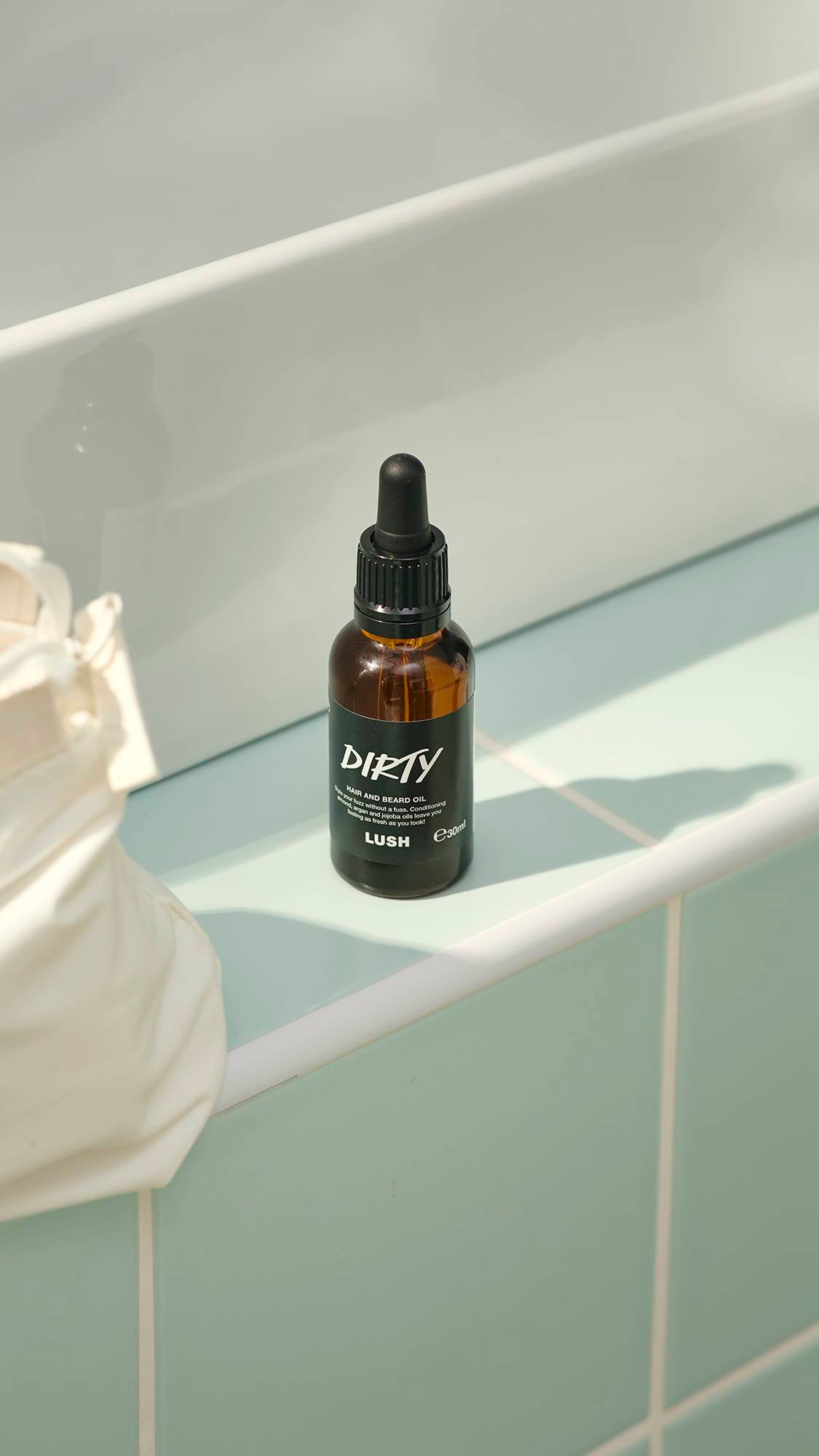 A small, amber glass bottle of "Dirty" hair and beard oil by Lush sits on a pale green tiled ledge. The bottle features a black label and a black pipette dropper lid. A white washbag is partially visible on the left.