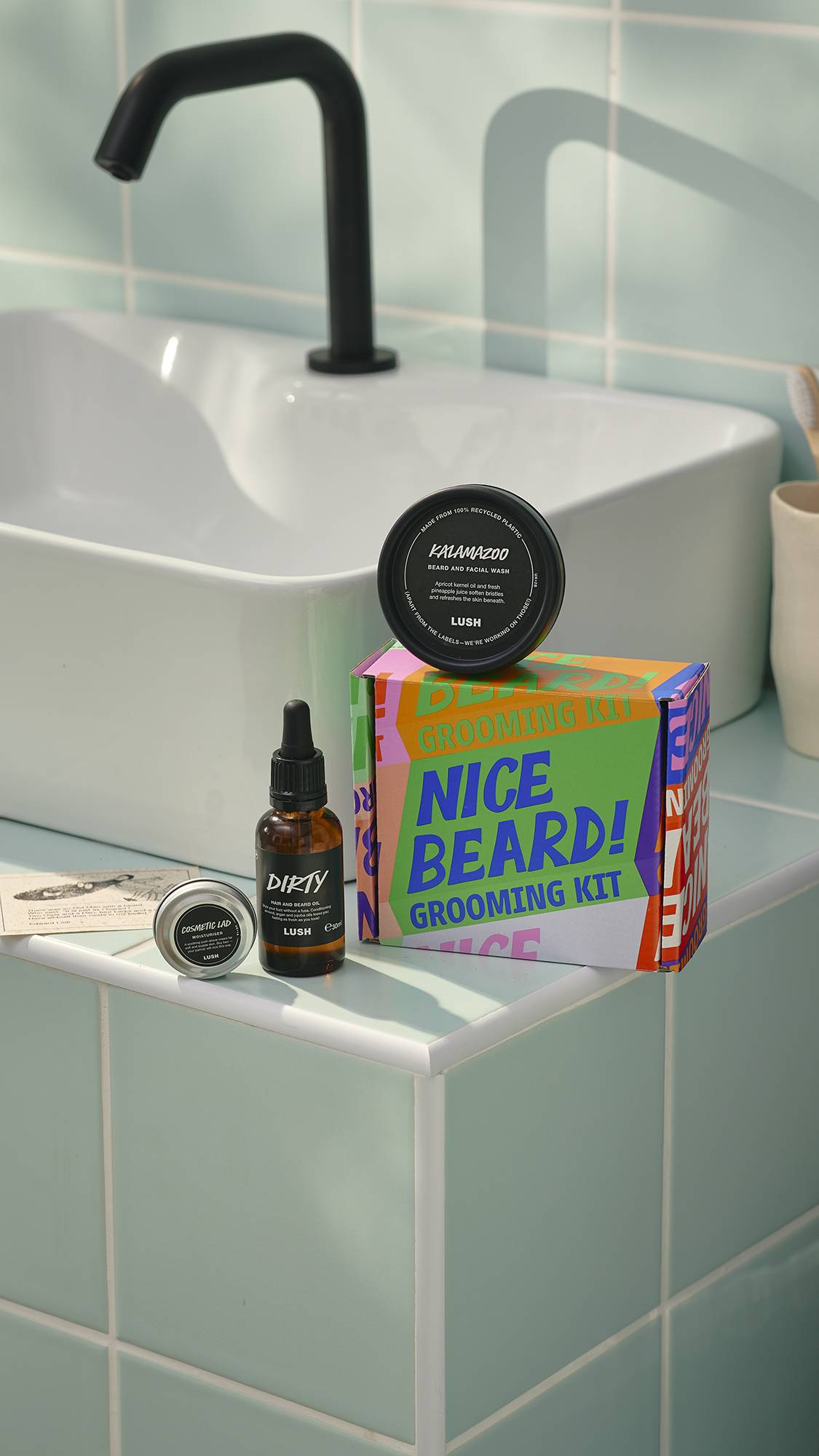 A Lush ‘Nice Beard!’ grooming kit sits on a tiled bathroom ledge next to a white sink. The set includes a colourful typographic box, a bottle of Dirty hair and beard oil, a tub of Kalamazoo beard wash, and a small tin of Cosmetic Lad moisturiser.