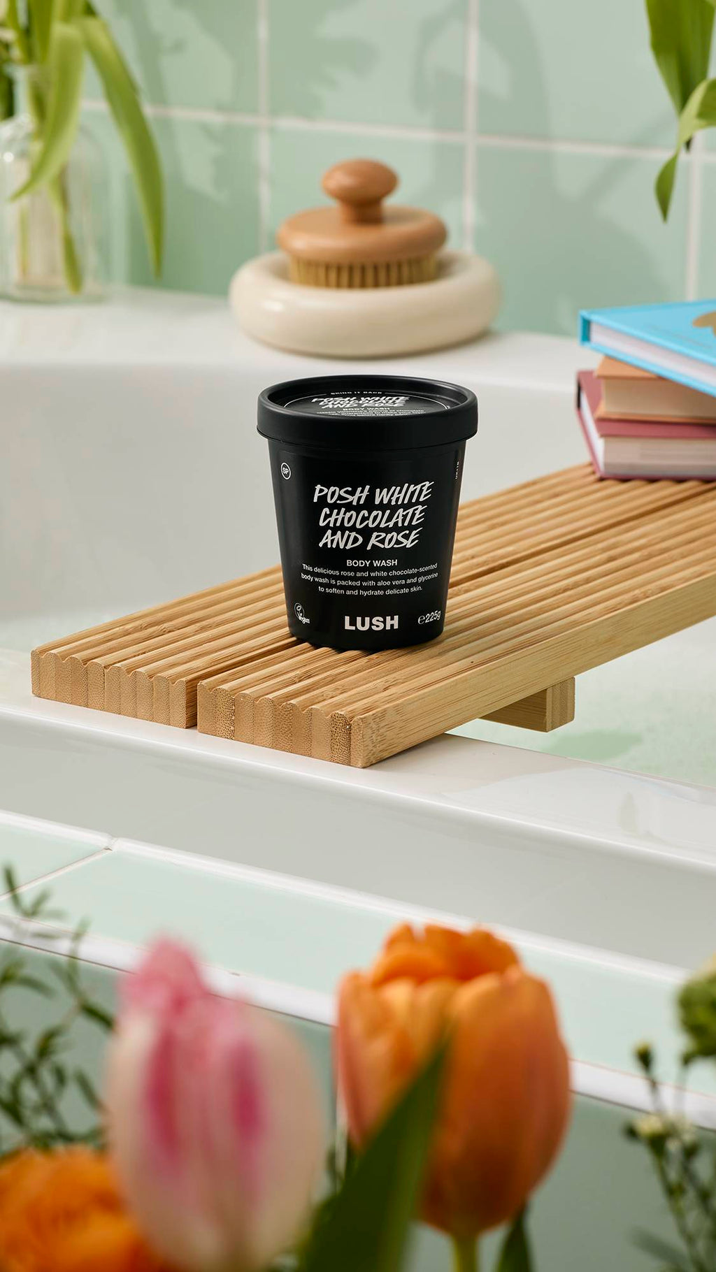 A black pot of "Posh White Chocolate and Rose" Body Wash sits on a wooden bath tray. The label notes it contains aloe vera and glycerine. The scene is set against mint green tiles with blurred tulips in the foreground.