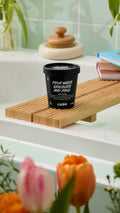 A black pot of "Posh White Chocolate and Rose" Body Wash sits on a wooden bath tray. The label notes it contains aloe vera and glycerine. The scene is set against mint green tiles with blurred tulips in the foreground.