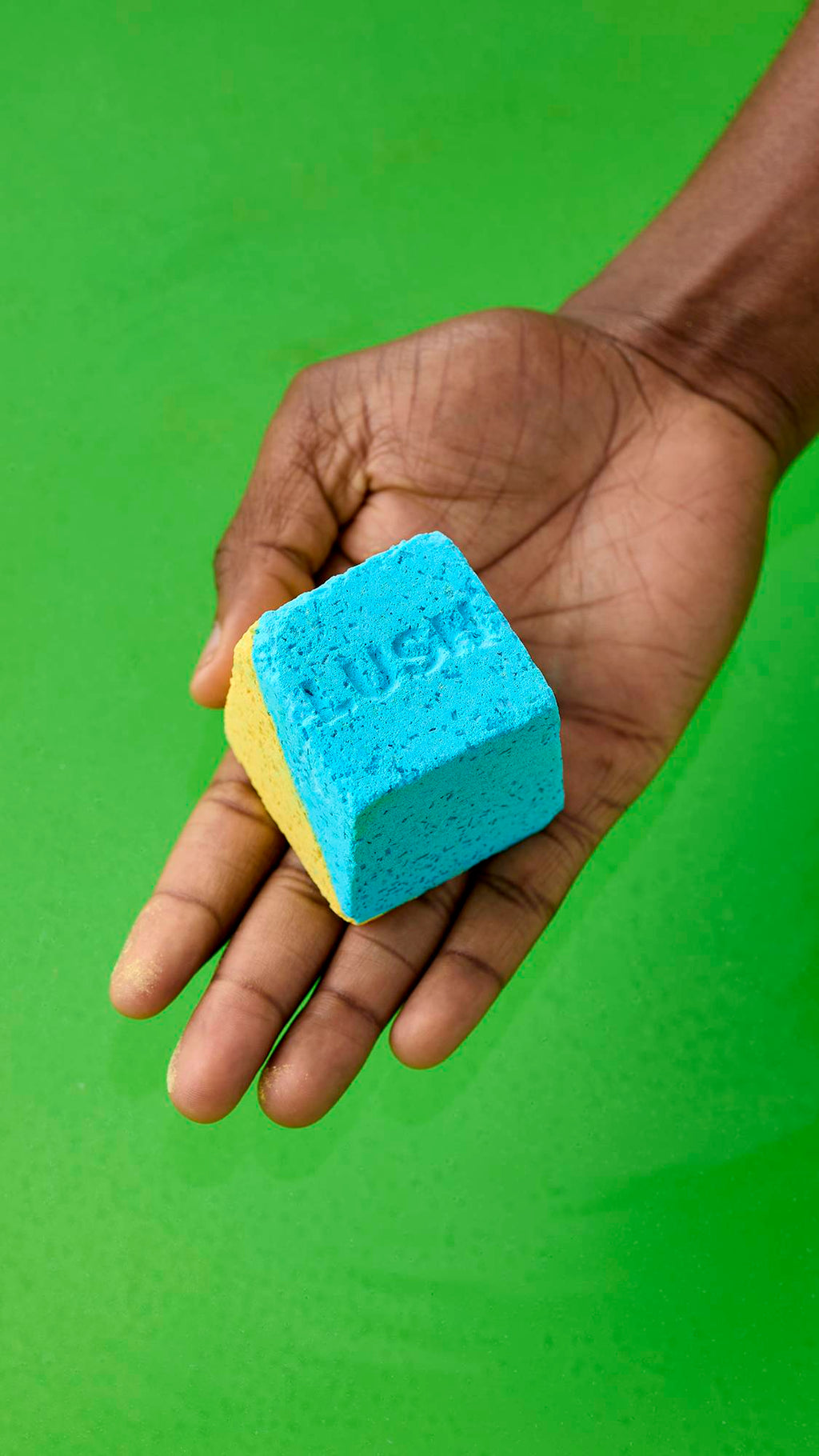 An open palm holds a cube-shaped, dual-coloured "Salt Water Soother" bath bomb in bright blue and yellow. The blue face has the word "LUSH" debossed on it, above deep green bathwater.