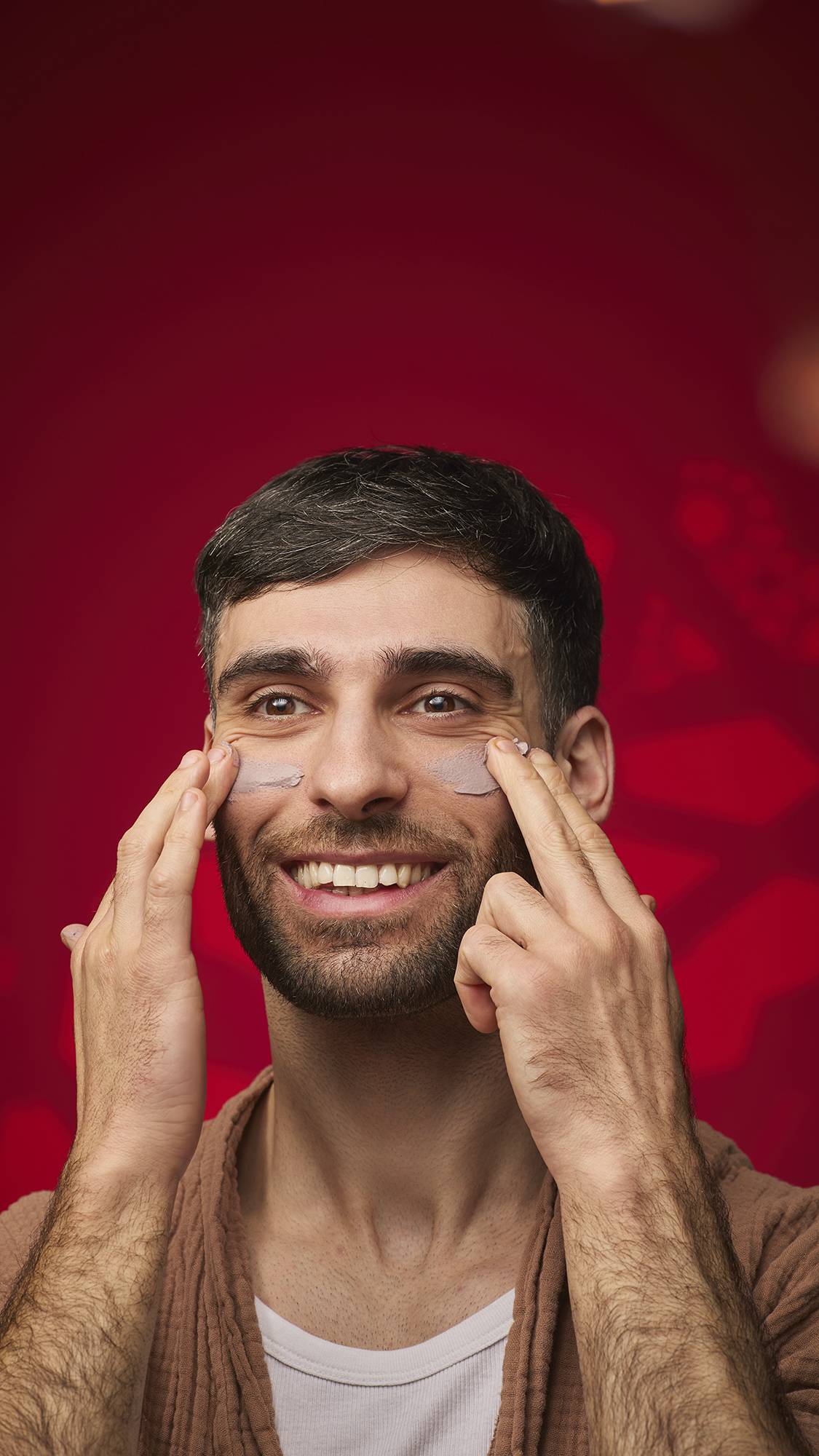A person smiles while applying dabs of the mauve Sipak face mask to their cheekbones. They wear a brown textured robe against a deep red background with soft golden bokeh lights.