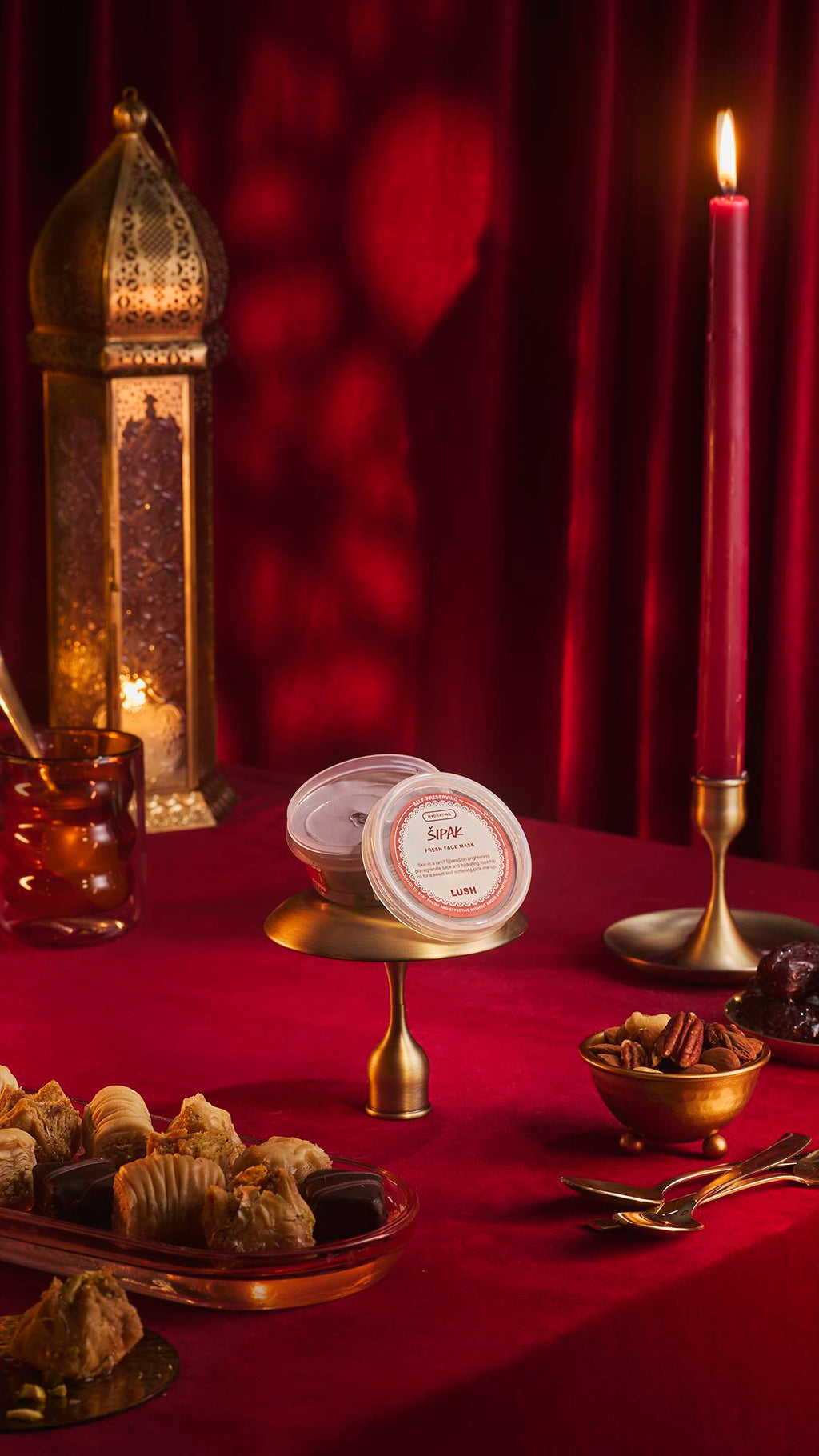 A pot of Lush 'Šipak' fresh face mask sits on a small brass stand atop a red velvet tablecloth. The festive scene features a tall gold lantern, a lit red candle, and bowls of nuts and pastries against a backdrop of draped red fabric.