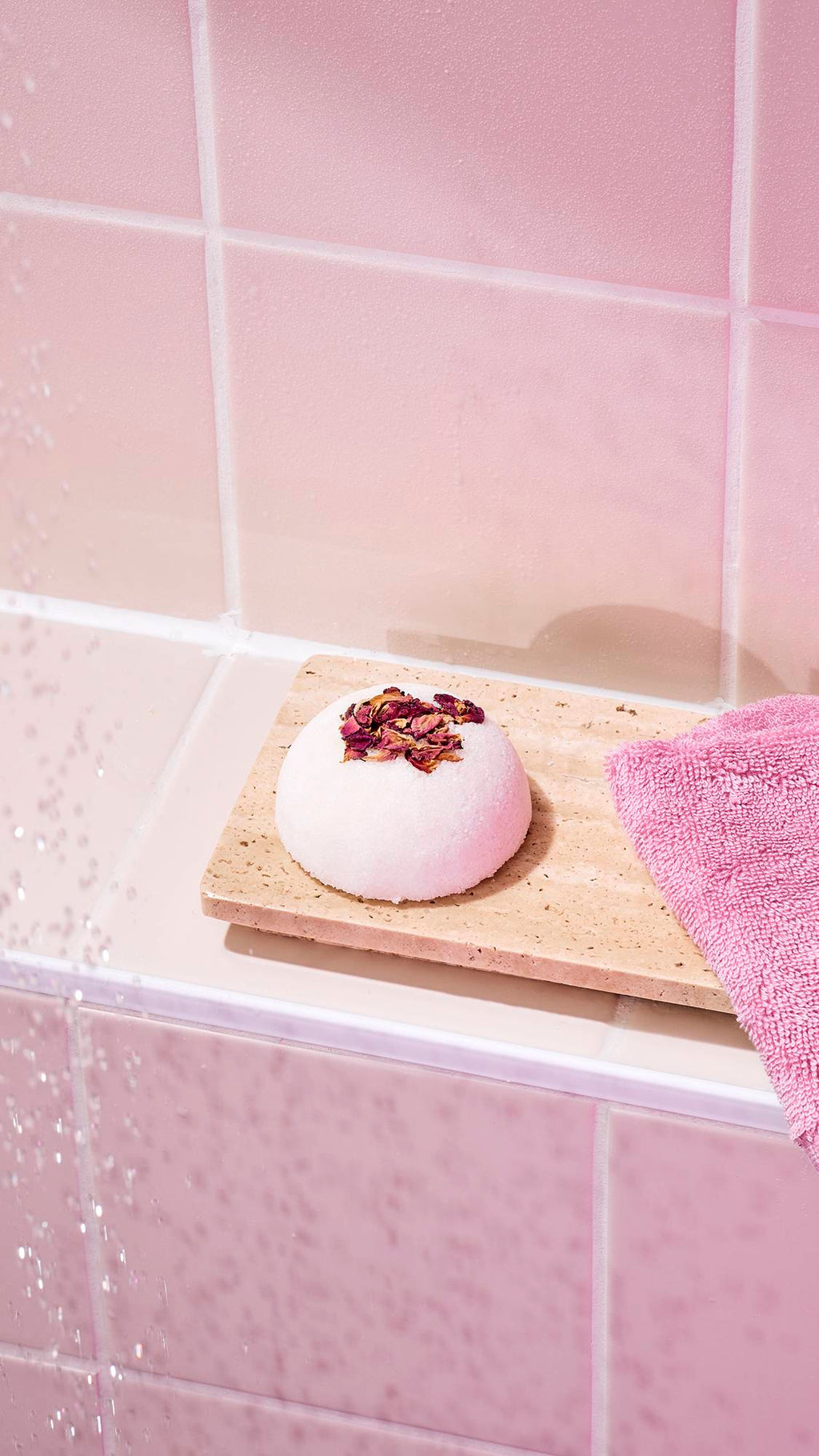 The image shows the "Comforter Rose" solid body scrub topped with dried rose petals, sitting on a beige stone tray in a bathroom. A pink flannel is tucked beside it, set against a background of soft pink wall tiles.