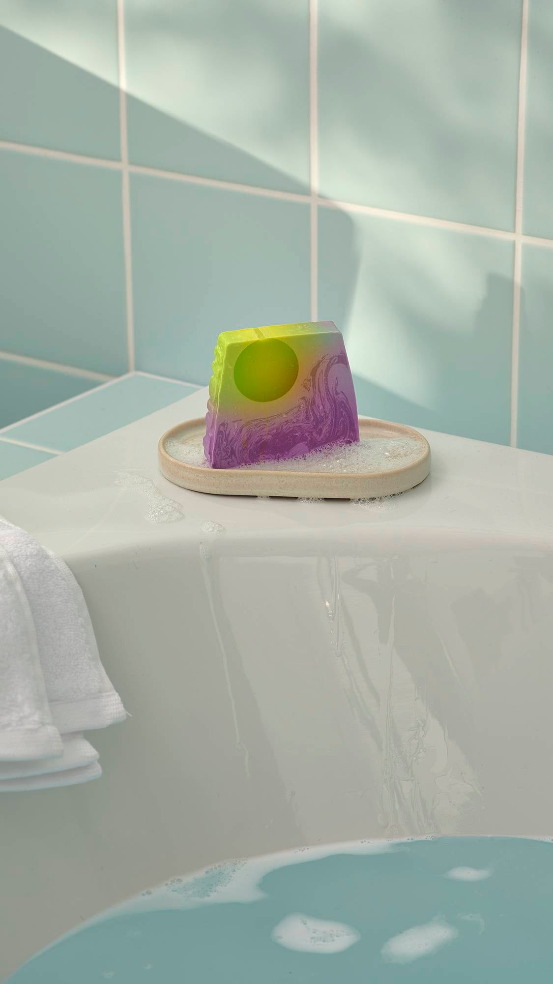The "To The Moon and Back" green and purple soap can be seen sitting in a small, oval dish on the corner of a white bathtub. The scene is set against mint green wall tiles, with a white towel visible on the left and soapy blue water in the bath.