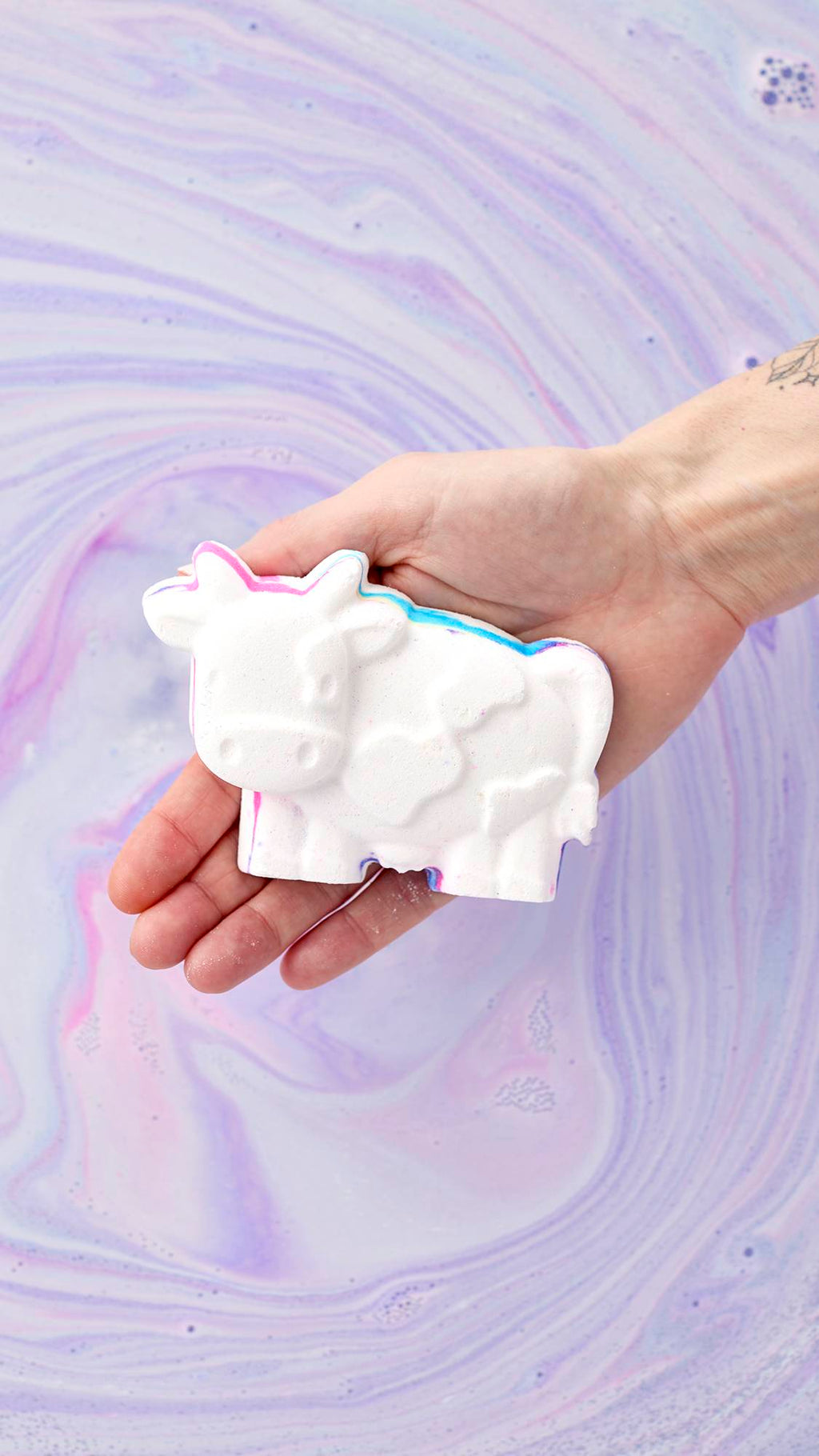A hand holds the white, cow-shaped Toby's Magic Cow Bath Bomb over a bathtub filled with swirls of pastel purple and blue water. This bath bomb features pops of hidden colors along its edges, ready to release a rainbow of foam into the tub.