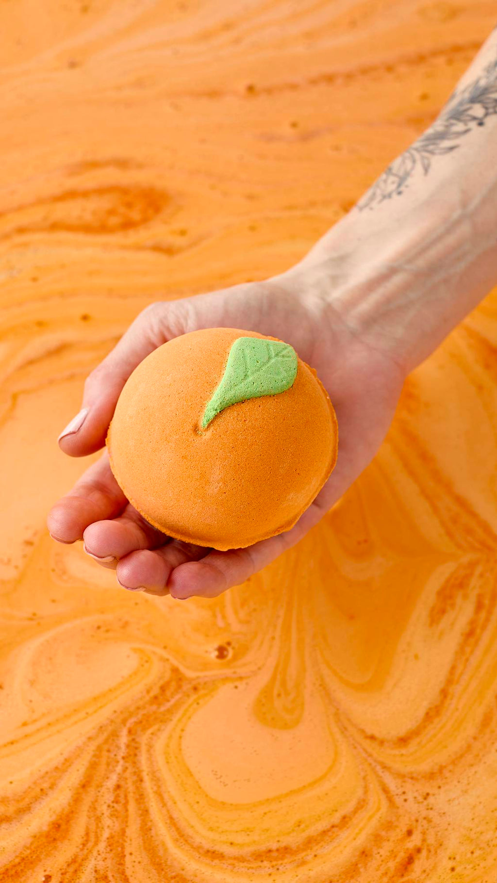 A hand holds the Peachy Bath Bomb over a tub of swirling orange water. The orange sphere, shaped like a real peach with a green leaf, creates a vibrant, citrusy soak.