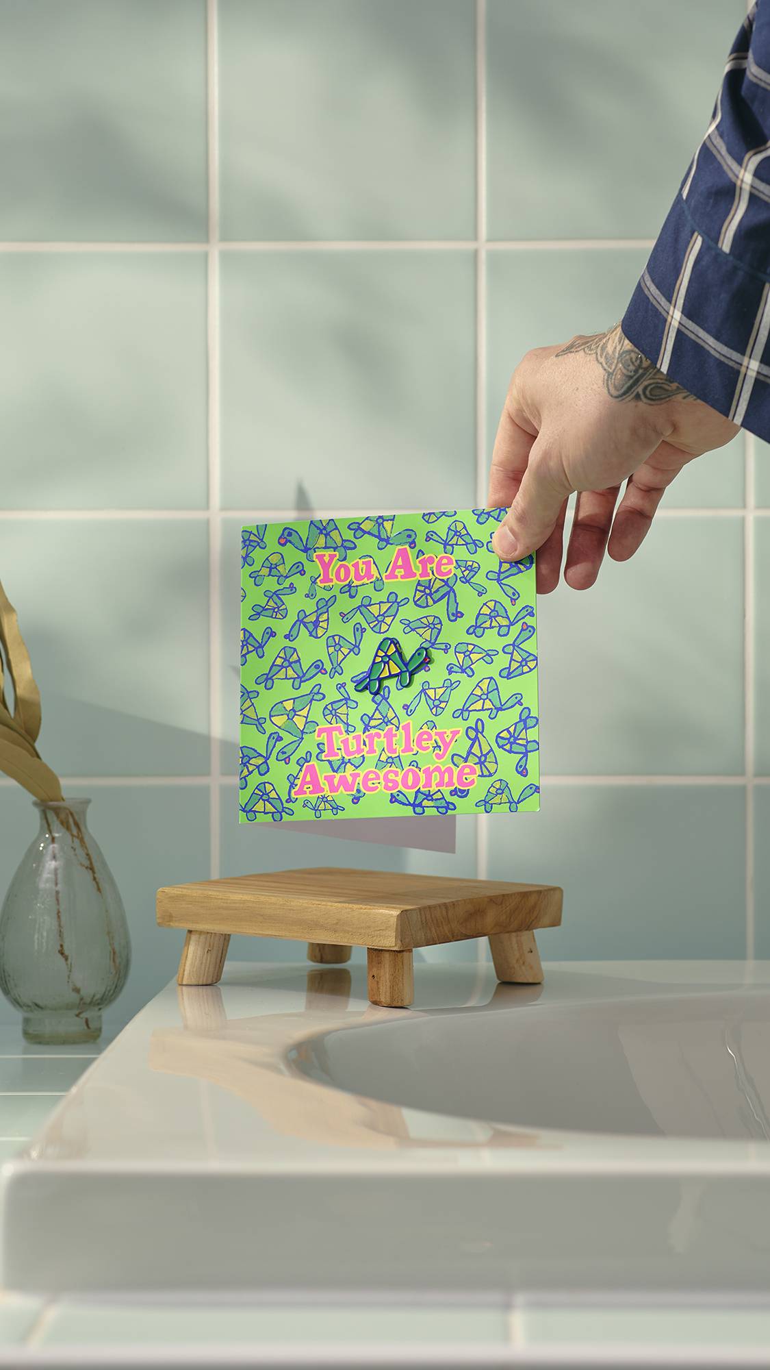 A tattooed hand holds a ‘Turtley Awesome’ greeting card over a wooden soap dish in a bathroom. The green card features a small turtle enamel pin and a repeating turtle pattern, set against a background of pale blue wall tiles.