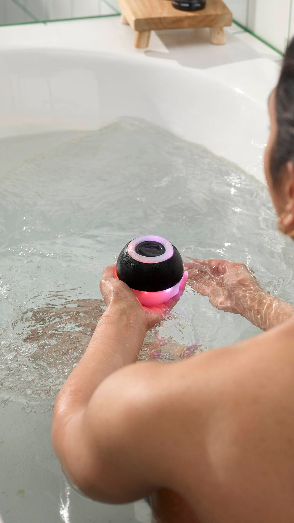 A model in a bathtub holds a black, spherical, bath bomb-shaped waterproof speaker that glows with pink and purple light. The device is partially submerged in the clear bathwater, with ripples expanding around the person's hands.