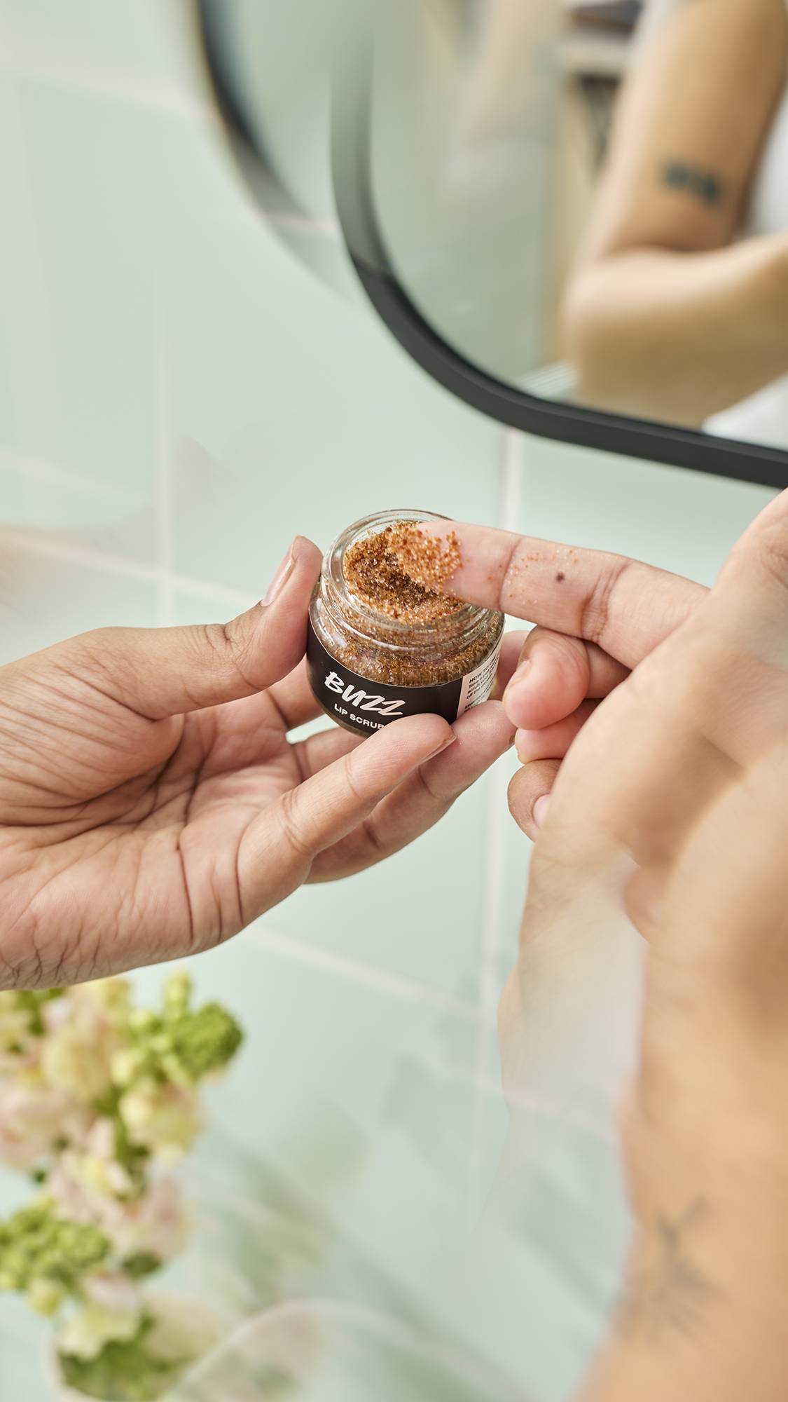 A close-up shot shows a model's finger dipping into a small glass jar of Lush "Buzz" Lip Scrub to scoop out the textured, caramel-coloured product. The scene is set against a background of pale green bathroom tiles and soft floral decorations.