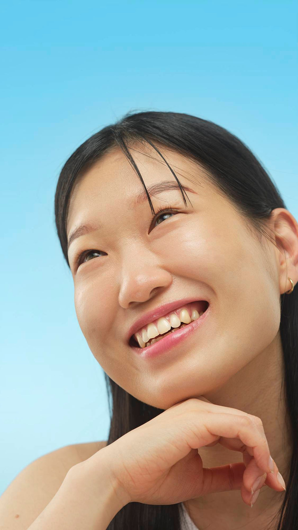 A close-up vertical portrait of a smiling person with dark hair, looking up and left. Their chin rests on their hand, and their face has a healthy, radiant sheen from the application of the Calm primer, set against a bright blue background.