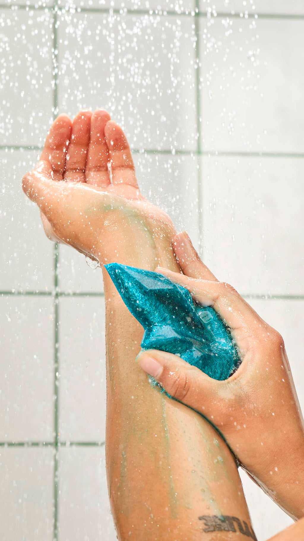 A hand rubs a shimmering teal, crystal-shaped shower gummy soap down a wet forearm under spraying water. White tiled walls with green grout are in the background.
