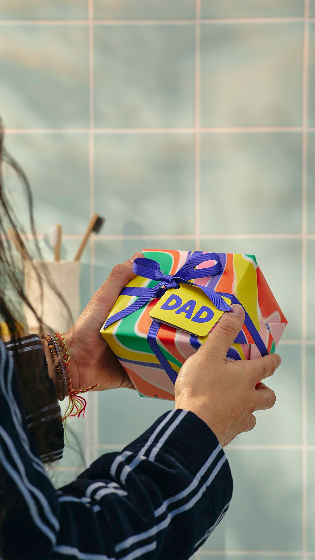 A close-up image of a model's hands holding a square gift wrapped in vibrant, swirling patterns of orange, yellow, and green. It is tied with a purple ribbon and a yellow tag that says 'DAD'. In the background are light blue bathroom tiles.