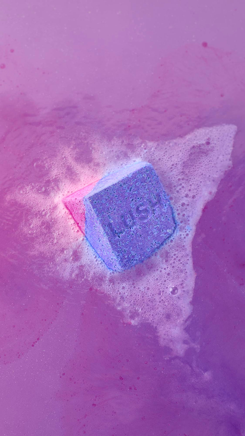 A top-down view of the Deep sleep Salt Cube bath bomb fizzing in vibrant purple water. The pink and purple cube is surrounded by a thick, frothy cloud of white bubbles and foam.