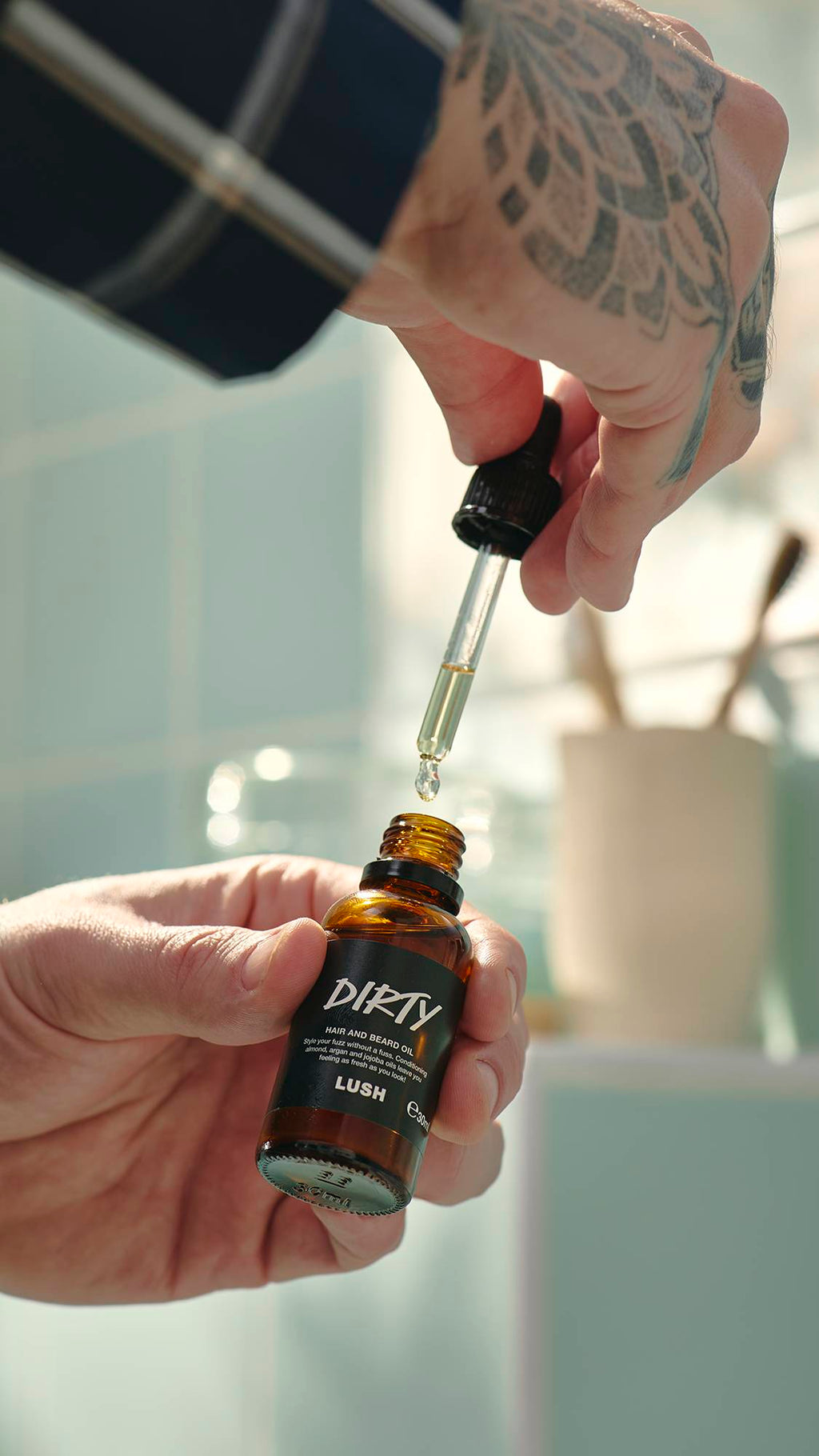 A model with a large geometric tattoo on their hand holds a small amber glass bottle of Lush "Dirty" hair and beard oil. They use a glass pipette to draw out a drop of the pale gold oil. Light green tiles are blurred in the background.