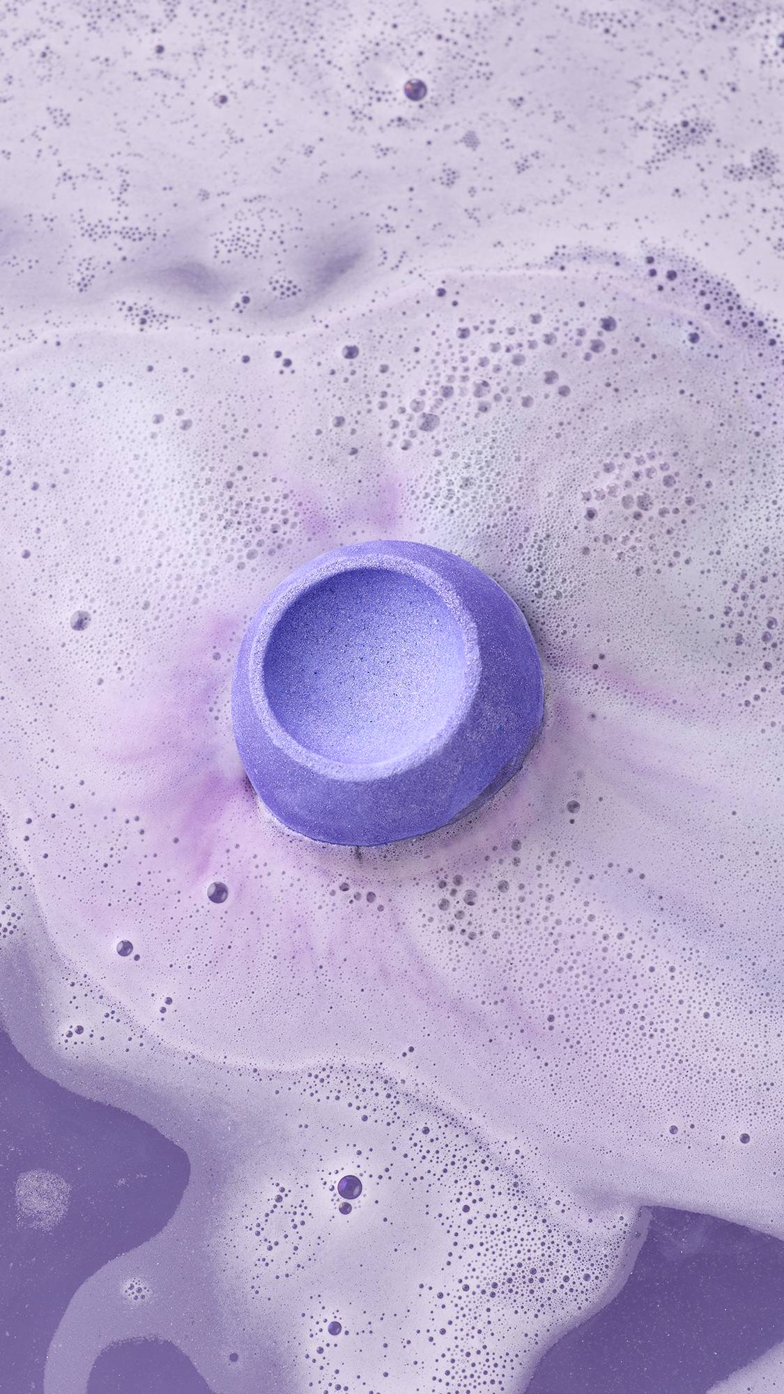 A vibrant purple Lush "Goddess" bath bomb fizzes in lilac-coloured water, creating a thick layer of white and pale purple foam. The bath bomb has a bowl-shaped indentation on top and is surrounded by delicate bubbles on the water's surface.