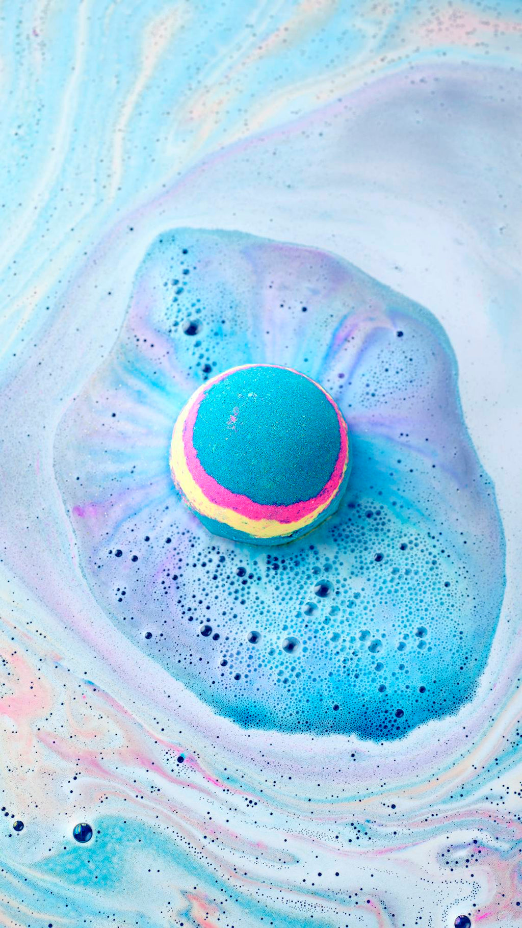 The Intergalactic bath bomb, with its blue, pink, and yellow stripes, floats in a bath. It creates a vibrant blue cloud of fizz on the surface, surrounded by swirling patterns of pastel blue, pink, and white foam.