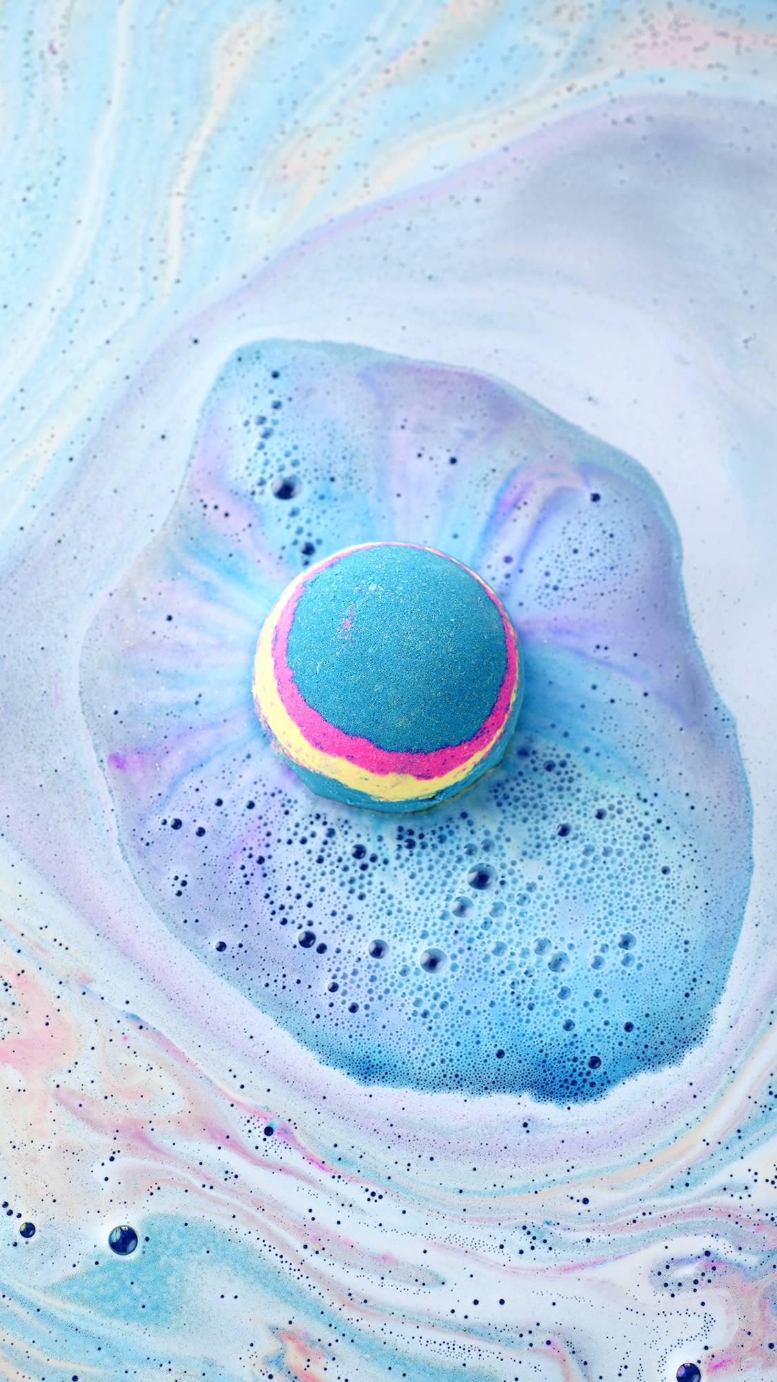 The Intergalactic bath bomb, with its blue, pink, and yellow stripes, floats in a bath. It creates a vibrant blue cloud of fizz on the surface, surrounded by swirling patterns of pastel blue, pink, and white foam.