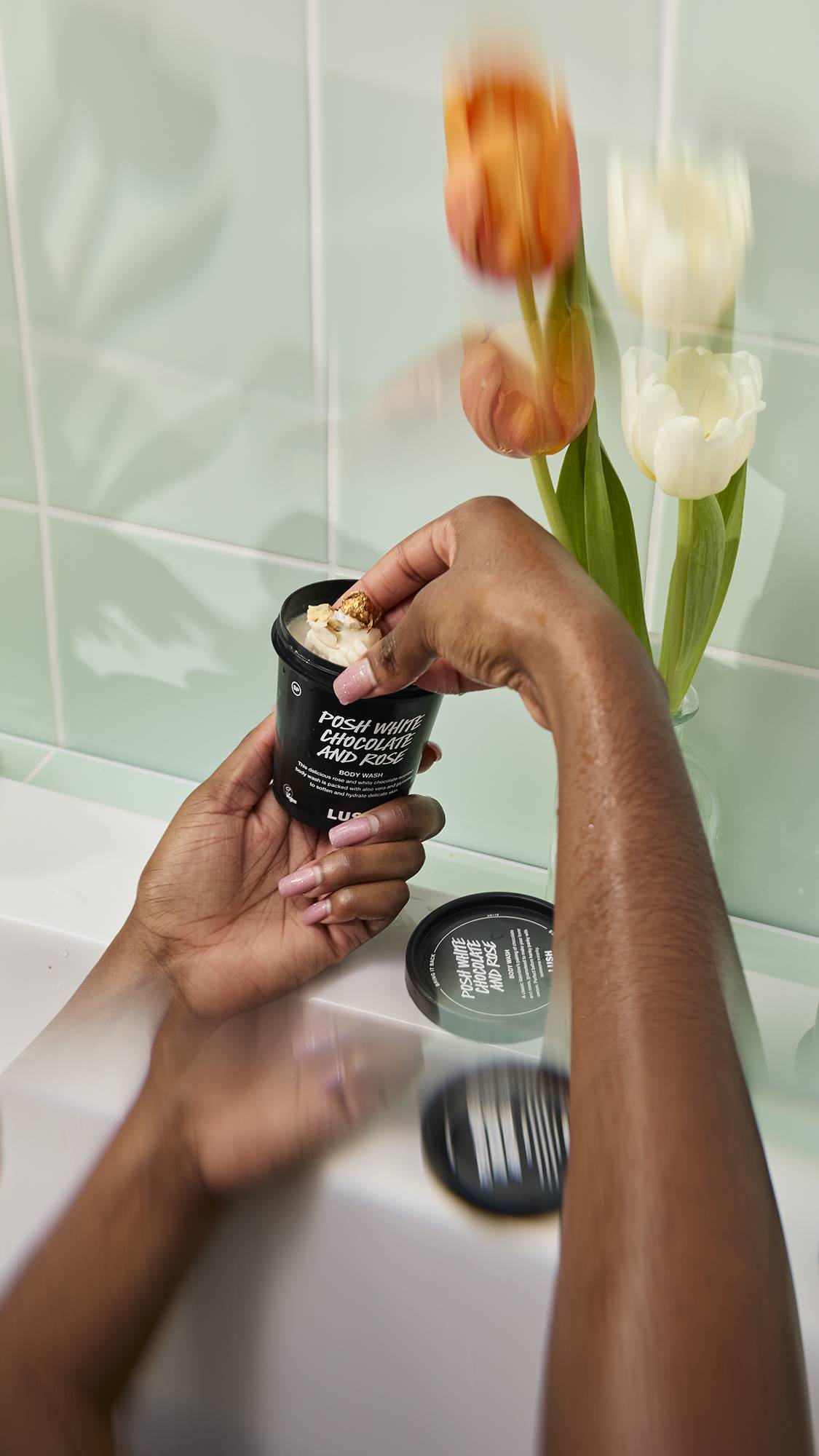 A person sitting in a bath scoops thick, white "Posh White Chocolate and Rose" Body Wash from a black pot. The scene features wet hands and blurred tulips against a mint green tiled background.