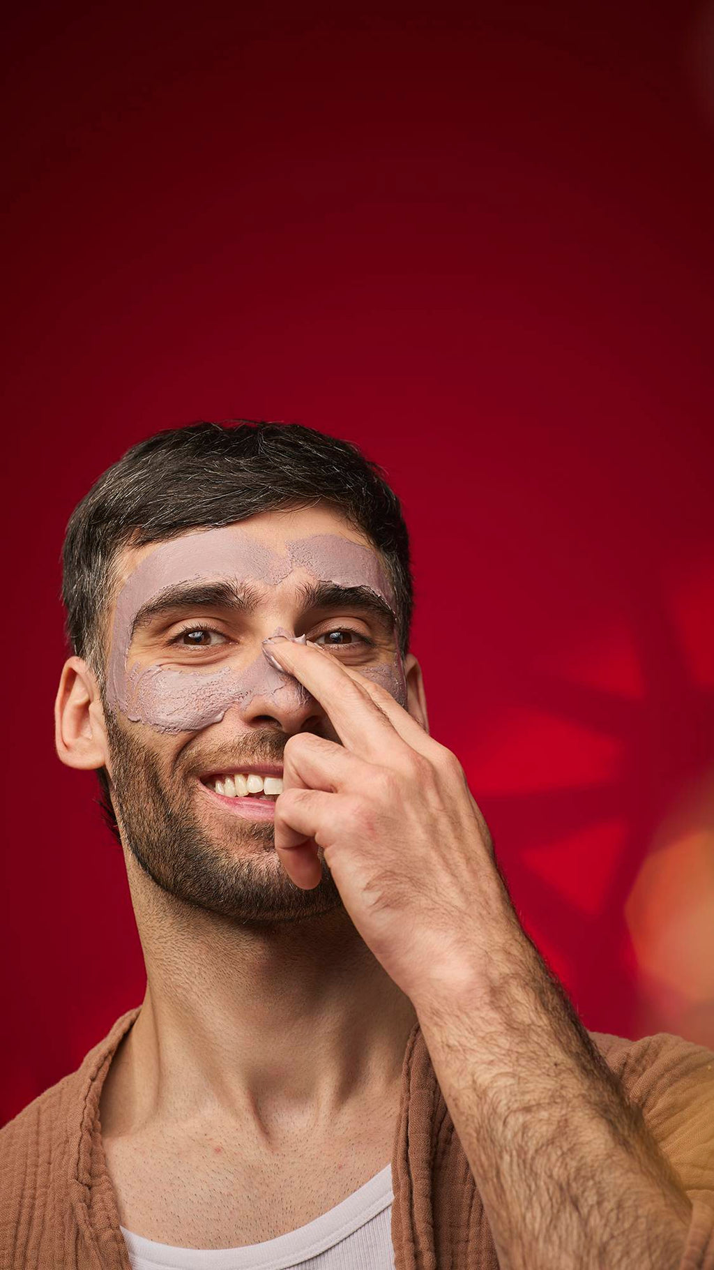 A person smiles while applying a thick, mauve-brown Sipak face mask to their nose and cheek area. They are wearing a brown robe against a deep red background with decorative bokeh patterns.