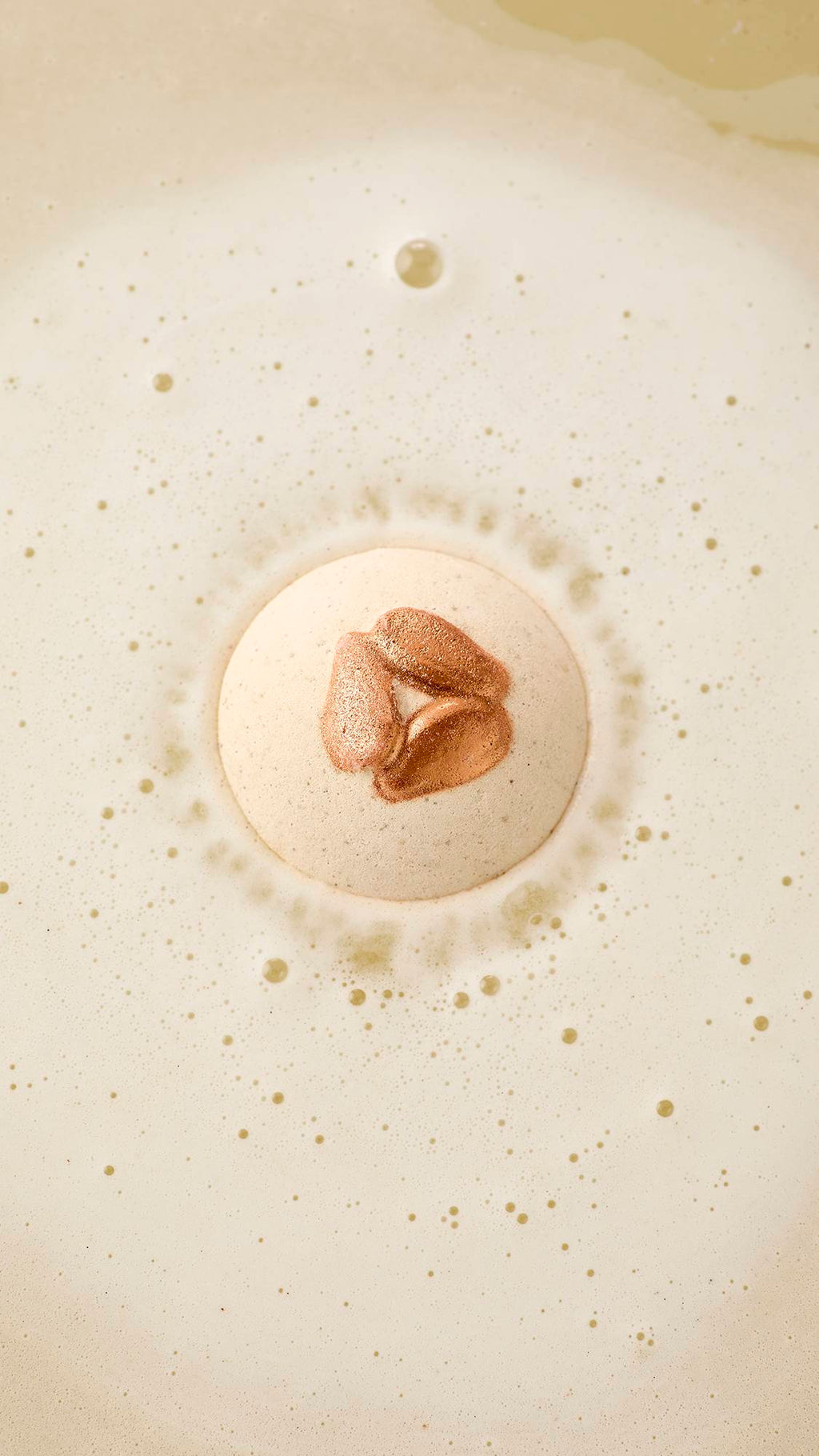 An aerial view of a speckled, off-white "Super Milk" bath bomb with 3 shimmering bronze, embossed almonds, as it begins to dissolve. It is surrounded by a ring of fine, frothy white bubbles against a creamy, pale-coloured water surface.