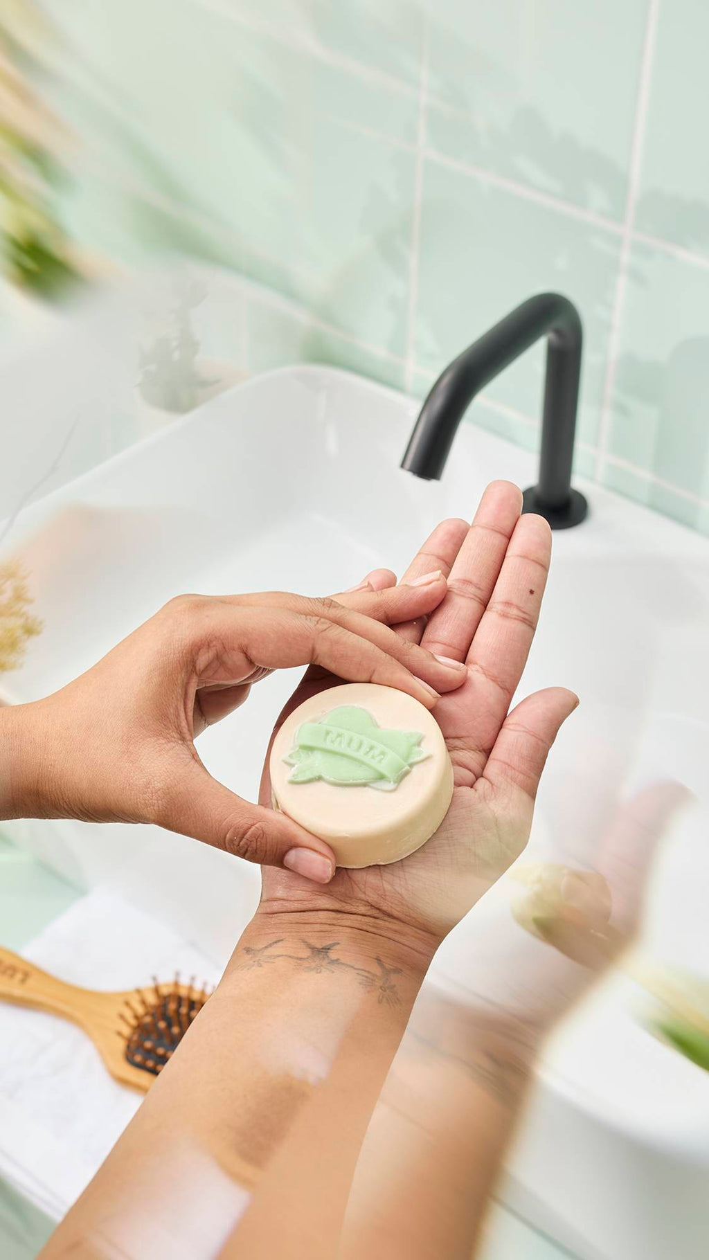 Hands hold a round, cream-coloured "Symbol of Love" body oil bar featuring a green heart and banner design reading "MUM" over a bathroom sink. A tattoo of three flying birds is visible on the wrist against a mint green tiled background.