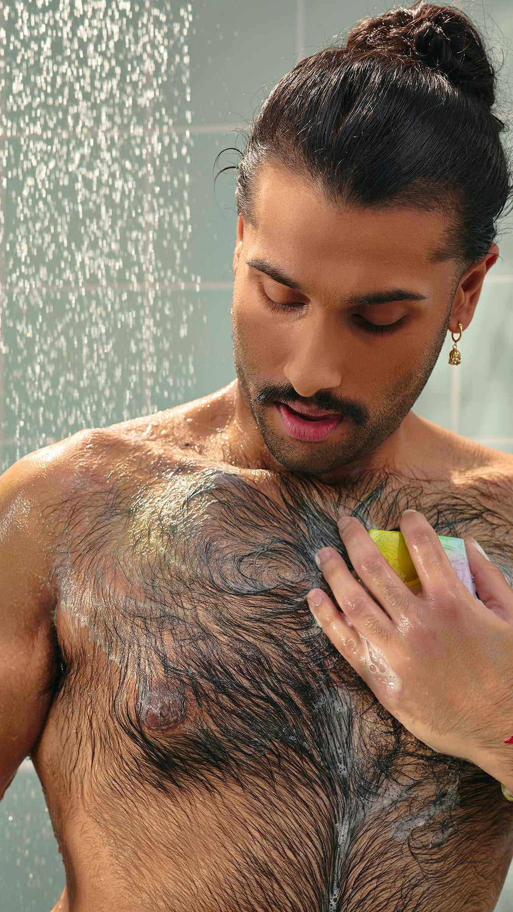 The image shows a model with dark hair in a bun. There is a running shower behind on pale green tiles. The model is using the "To The Moon and Back" soap slice to lather up their chest.