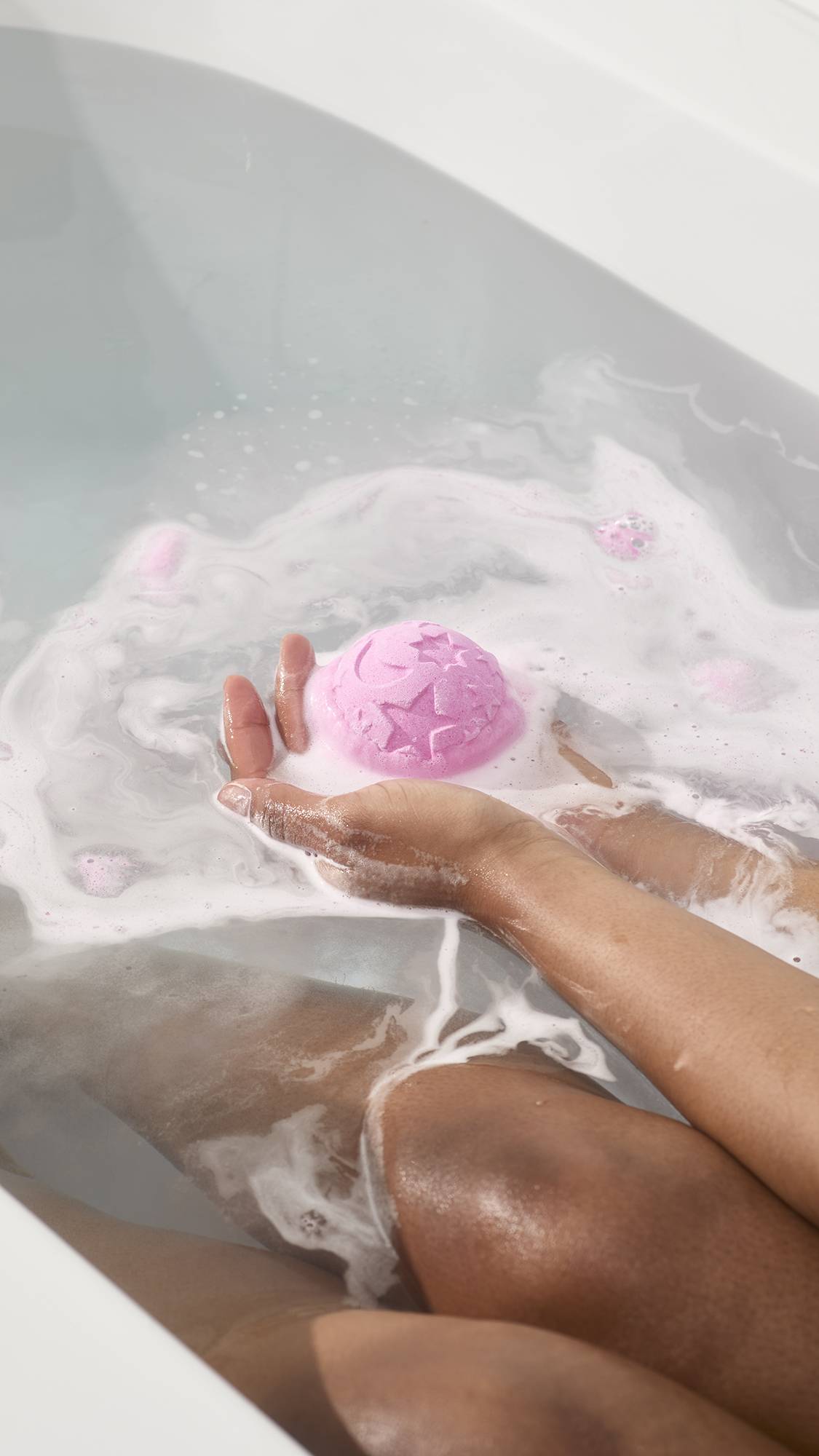 Lavender Twilight Bath Bomb with star and moon designs fizzes in hands over white bathwater foam.