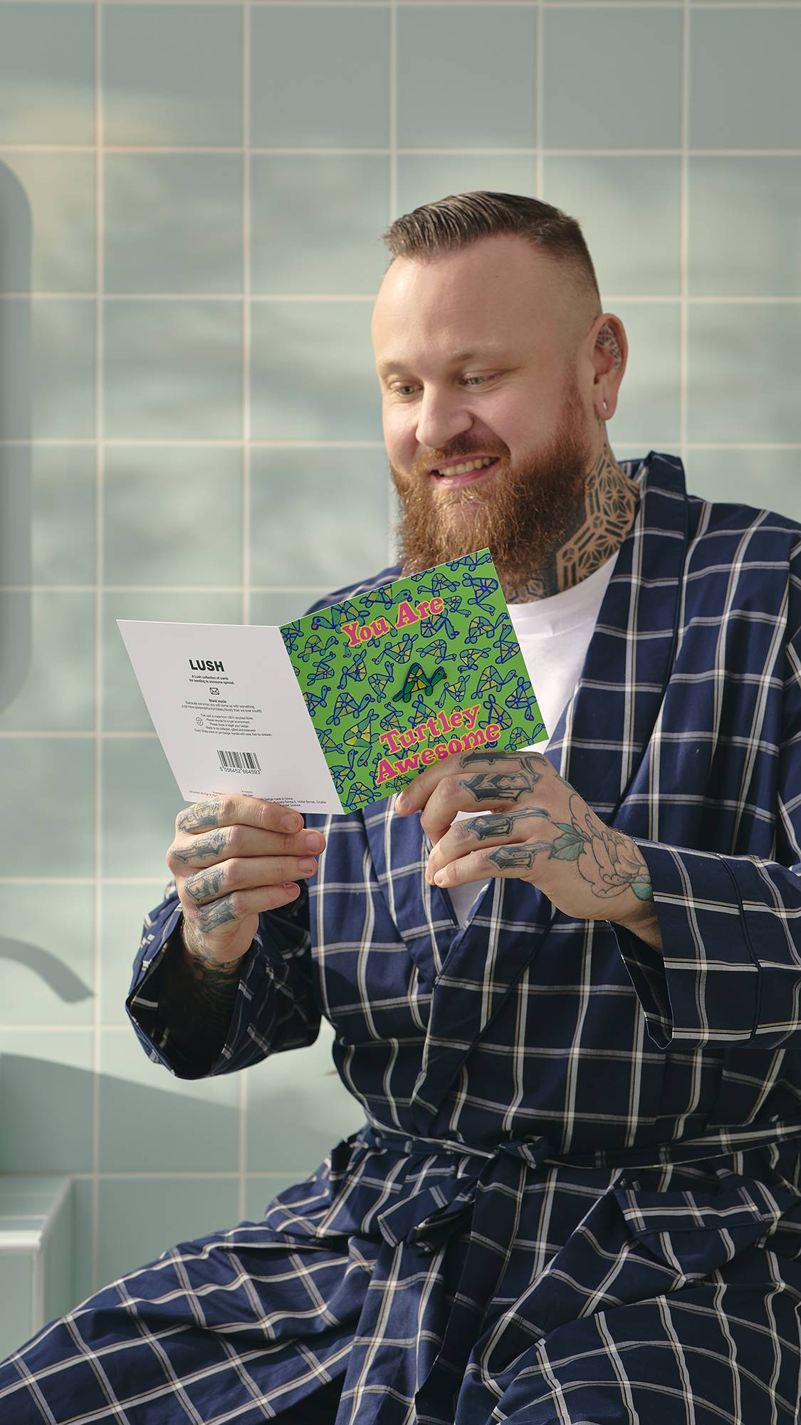 A model with a beard and tattoos, wearing a blue checked dressing gown, smiles while reading the inside of a 'Turtley Awesome' card, with a removable enamel turtle pin badge. They are in a bathroom with pale blue tiles in the background.