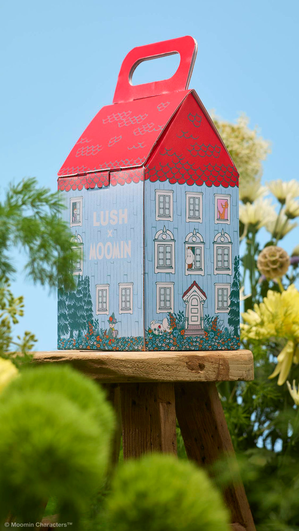 A house-shaped "LUSH x MOOMIN" gift box sits on a small wooden stool. The box is illustrated to look like a blue house with a red roof. Green foliage and yellow flowers frame the stool against a clear blue sky.