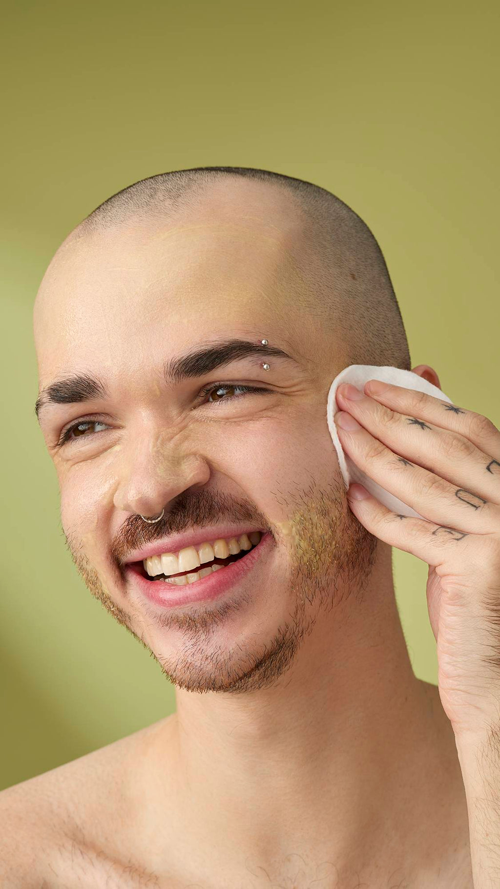 A person with a shaved head and facial piercings smiles while using a white cotton pad to wipe the Acai And Sacha Inchi cleanser from their cheek. They have tattooed hands and are shown against a solid olive-green background.