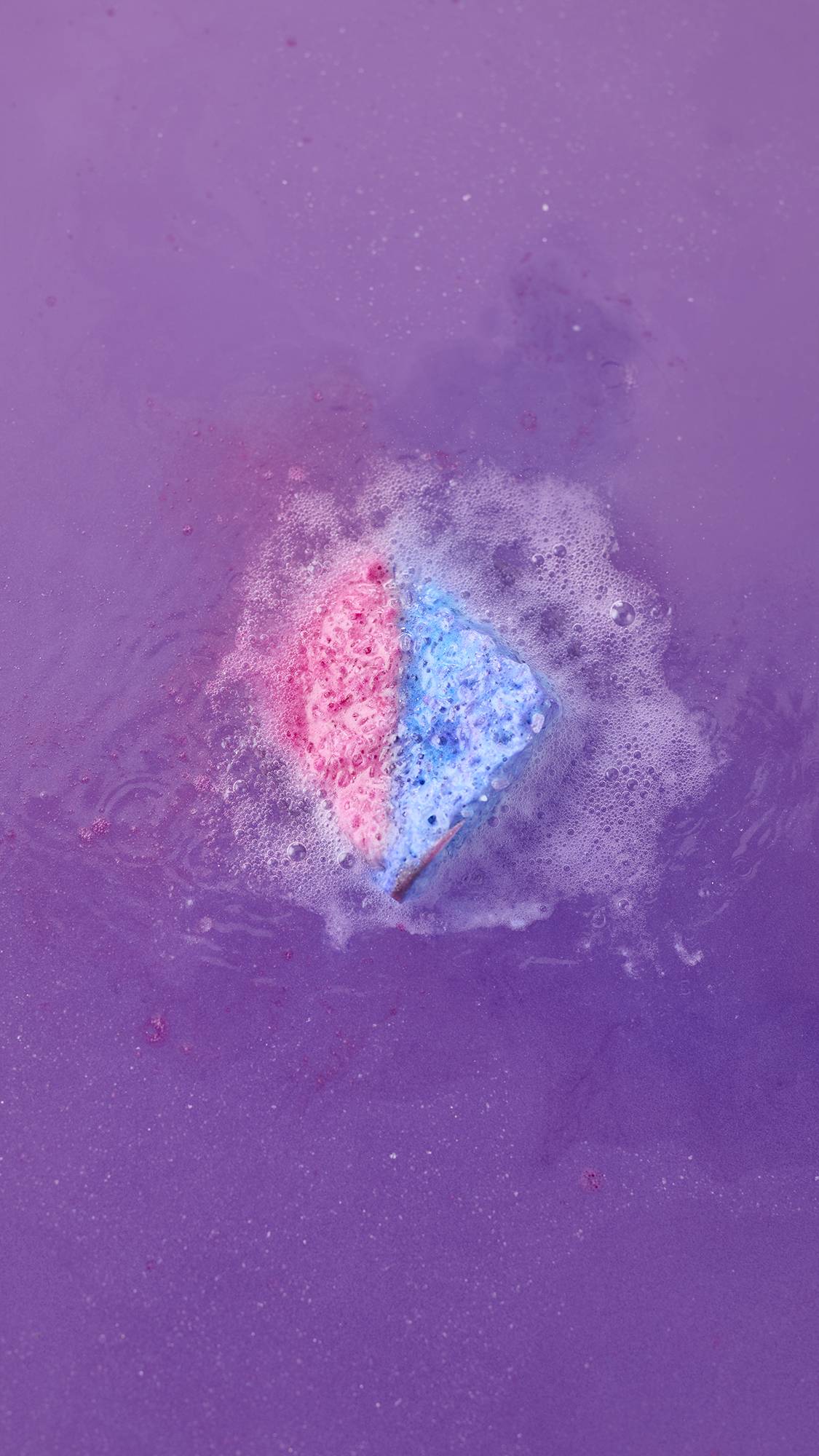 A top-down view of the Deep sleep Salt Cube bath bomb fizzing in deep purple water. The pink and blue cube is surrounded by a ring of frothy white foam and bubbles as it dissolves.