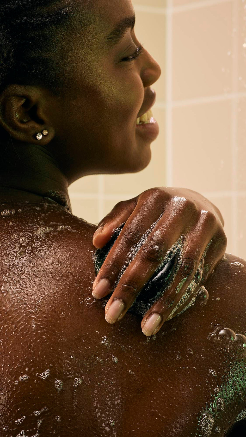 A model with braided hair uses a dark green, translucent "The Good Hour" shower jelly to wash her shoulder. Water from the shower streams over her skin and the jelly, creating a glistening effect against the backdrop of neutral-toned bathroom tiles.