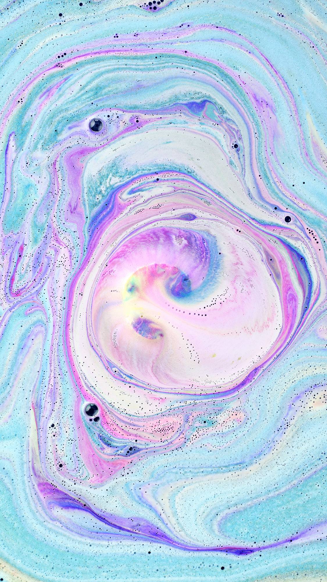 A top-down view of bathwater featuring a mesmerizing swirl of vibrant neon pink, bright blue, and soft white foam. The colors create a cosmic, marble-like pattern with tiny dark speckles scattered throughout the water's surface.