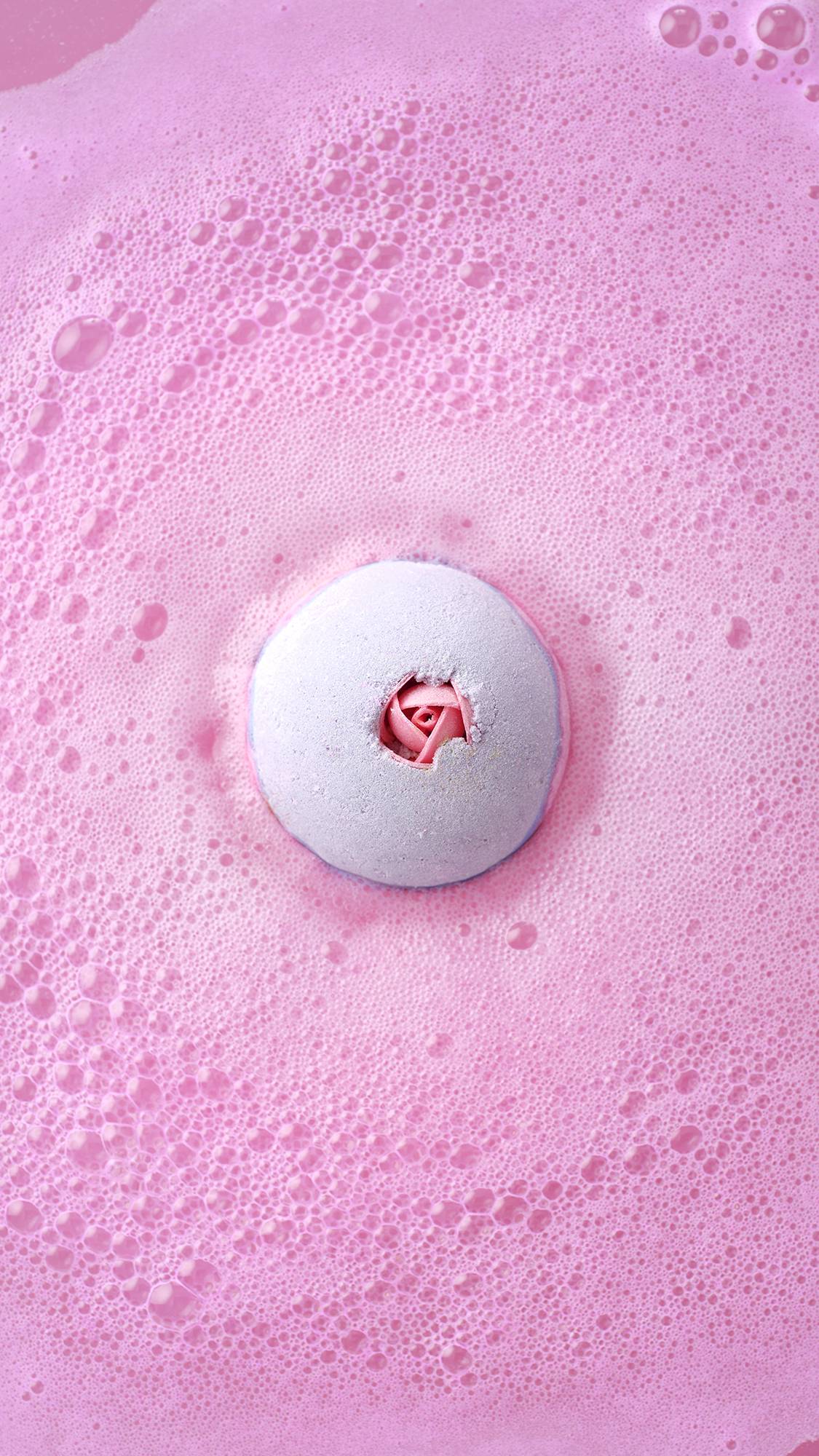 Top-down view of the Sex Bomb Bath Bomb dissolving in a sea of vibrant, frothy pink bubbles, with its signature rosebud detail visible in the center.