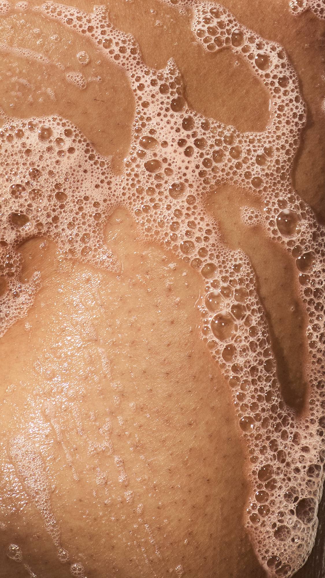 A close-up of wet skin covered in thick trails of soapy lather and clusters of small bubbles.