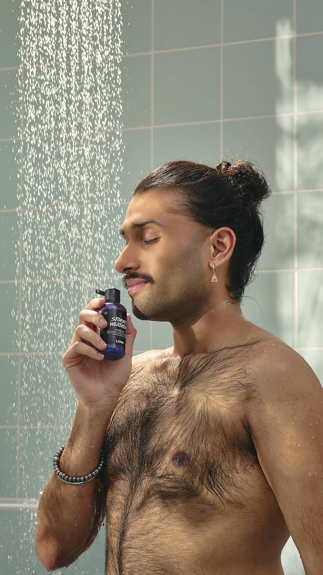 A model with dark hair in a bun and a moustache stands in a shower, holding a bottle of purple "Stormy Weather" shower gel to their nose. They have their eyes closed with a peaceful expression as water falls around them against pale green tiles.