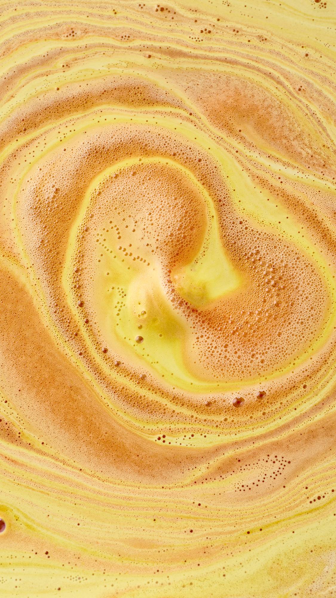 AN aerial, abstract view of swirling bath art created by the "Sweet As Honey" bath bomb. Vibrant streaks of amber-orange foam and pale yellow liquid spiral together, creating a marbled, creamy texture with tiny bubbles across the surface.
