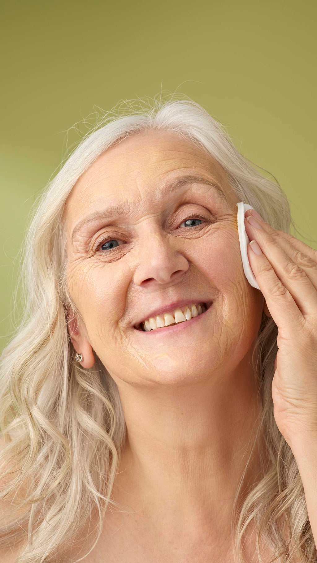 A model with long grey hair smiles while wiping the yellow Turmeric And Argan cleanser with a cotton pad. She is shown from the shoulders up against a solid olive-green background, highlighting the creamy texture of the facial product.