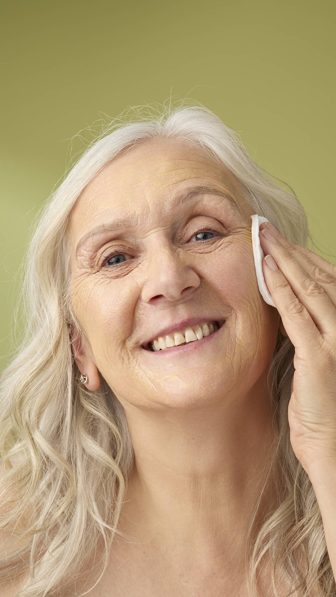 A model with long grey hair smiles while wiping the yellow Turmeric And Argan cleanser with a cotton pad. She is shown from the shoulders up against a solid olive-green background, highlighting the creamy texture of the facial product.