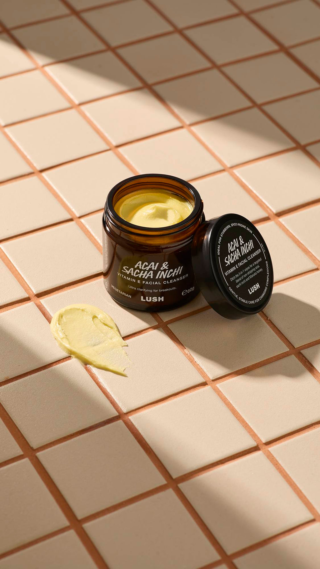 An open brown glass jar of Lush "Acai & Sacha Inchi" Vitamin E facial cleanser sits on a beige tiled surface. A thick dab of the pale yellow, creamy product is smeared on the tiles next to the jar and its black lid.