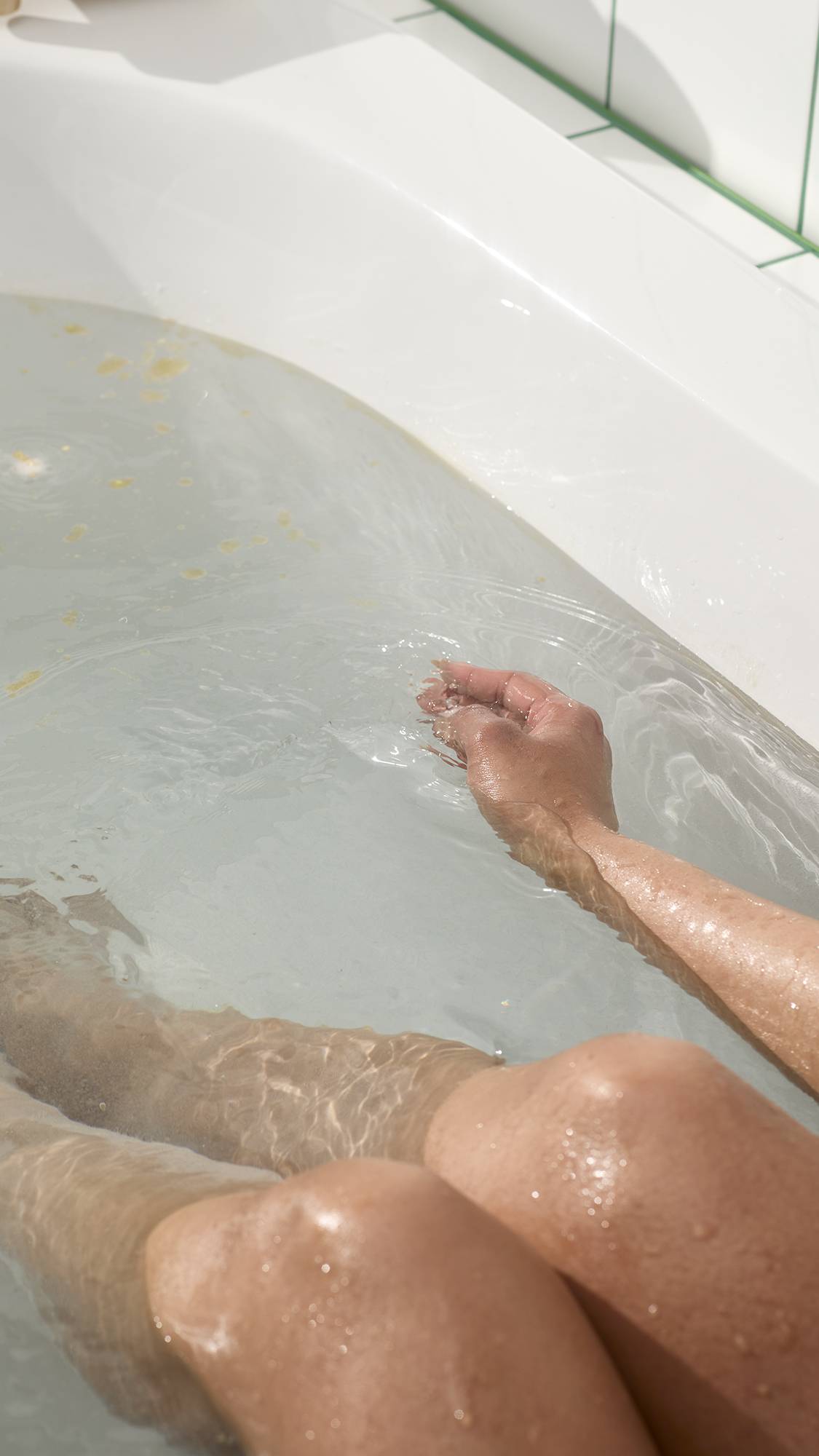 An aerial view of a person with their head tilted back and eyes closed, soaking in a bath of light blue water. The surface is covered in a thick layer of creamy, white foam and visible cocoa butter flecks from the Butterball bath bomb.