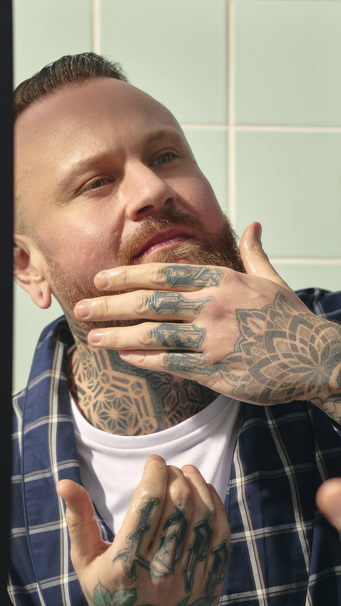 A model with a ginger beard and a blue-and-white checked dressing gown looks into a bathroom mirror. They have extensive tattoos on their neck and hands, and are using their fingers to massage the "Dirty" Hair and beard oil in.