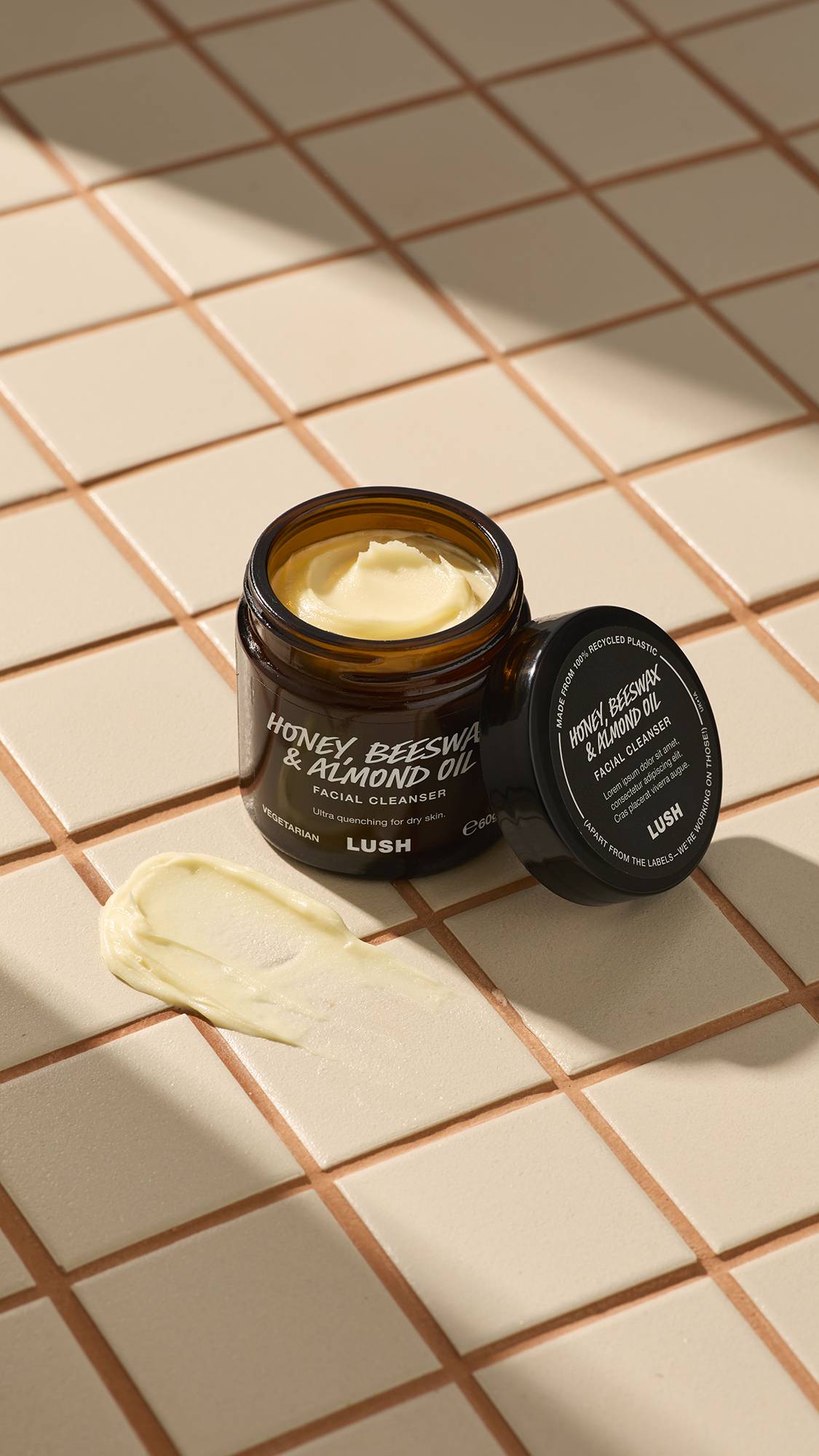 An open brown glass jar of Lush "Honey, Beeswax And Almond Oil" facial cleanser sits on a beige tiled surface. A thick dab of the pale yellow, creamy product is smeared on the tiles next to the jar and its black lid.